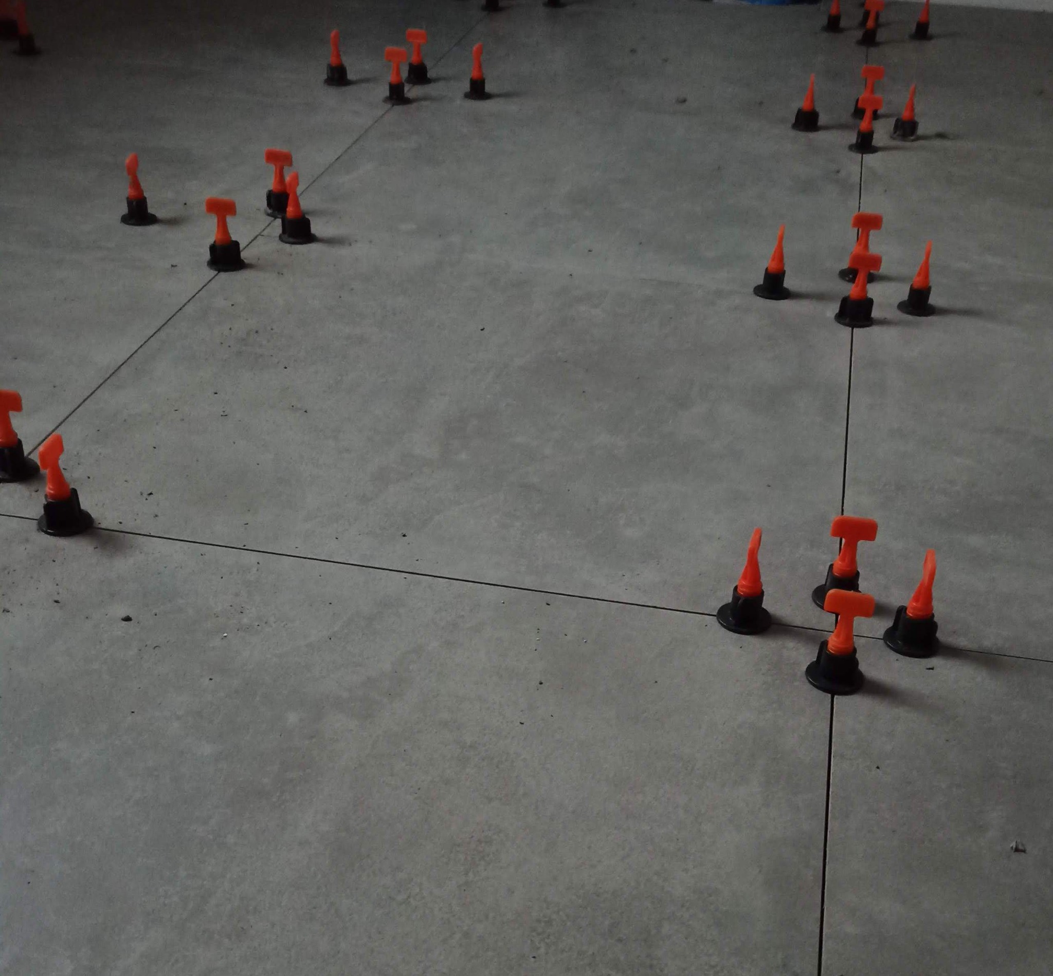 Szare płytki podłogowe z pomarańczowymi klipsami poziomującymi i czarnymi podstawkami, ułożone w rzędach z czarnymi liniami wyznaczającymi spoiny.