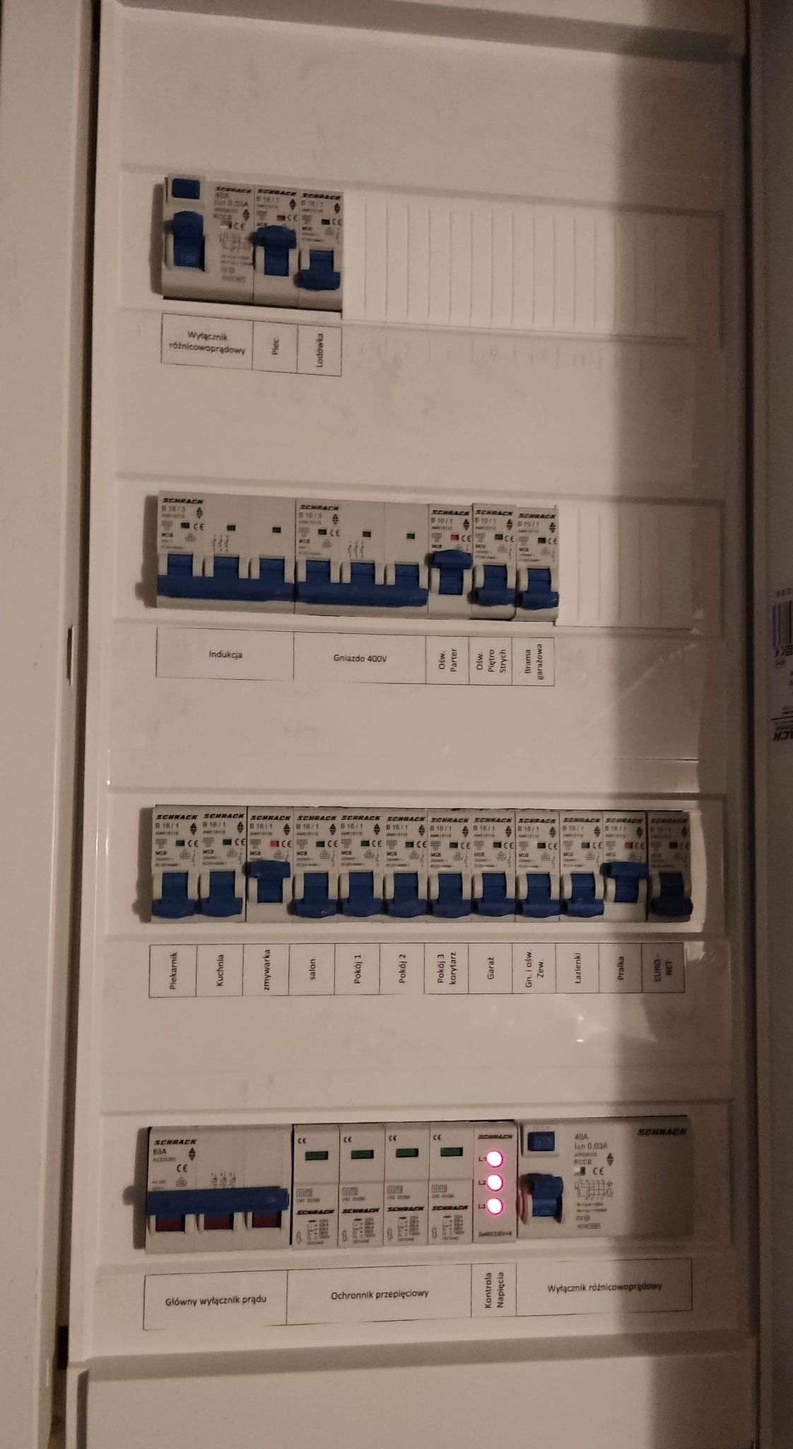 Rozdzielnica elektryczna z bezpiecznikami Schrack, opisana dla różnych obwodów: piekarnik, indukcja, gniazdo 400V, oświetlenie, kuchnia, zmywarka, salon, pokoje, garaż, łazienka.