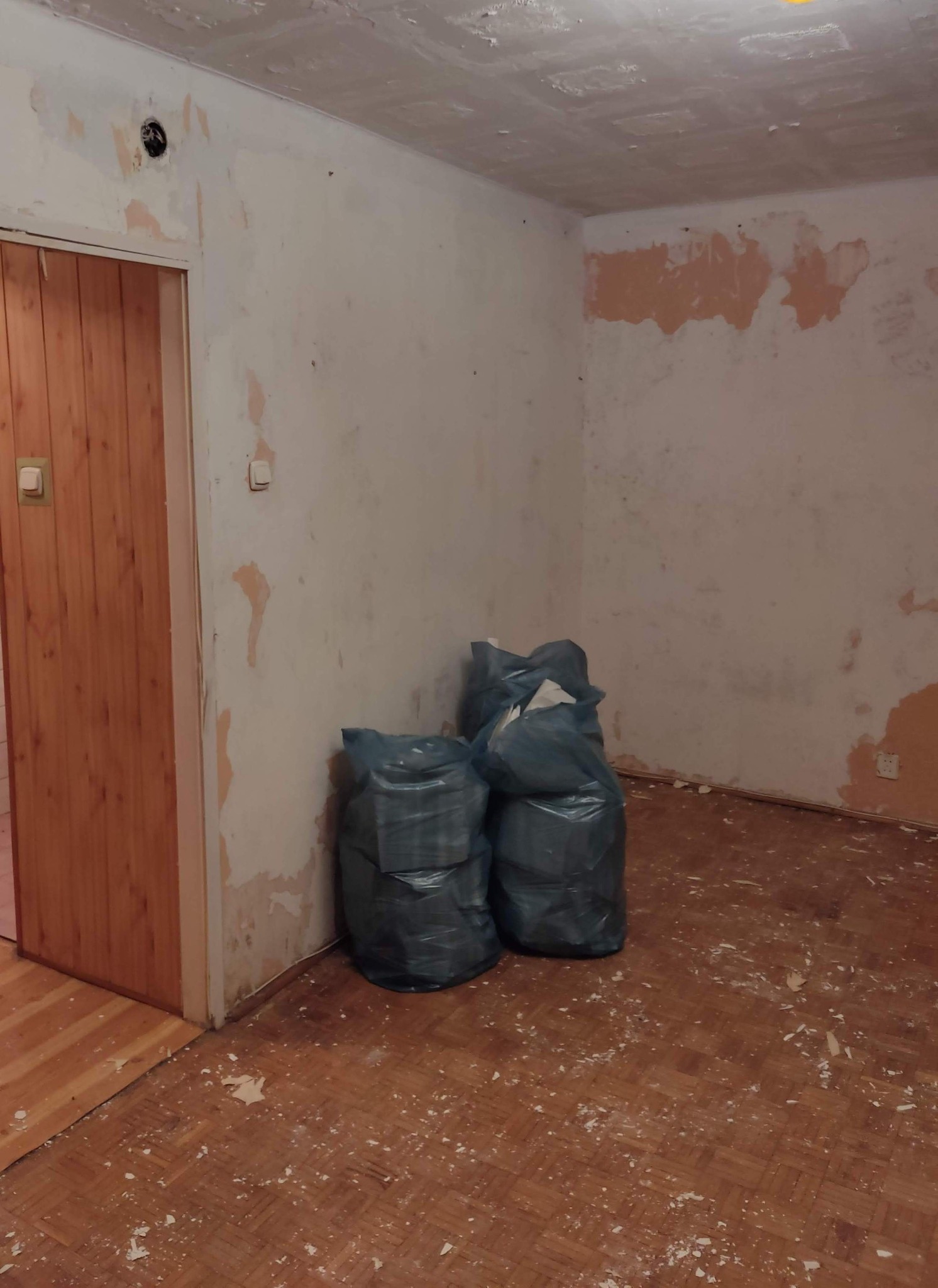 Pokój w trakcie demontażu: ściany z resztkami farby, podłoga z resztkami gruzu, worki z odpadami. Widoczne ślady po remoncie, przygotowanie do dalszych prac.