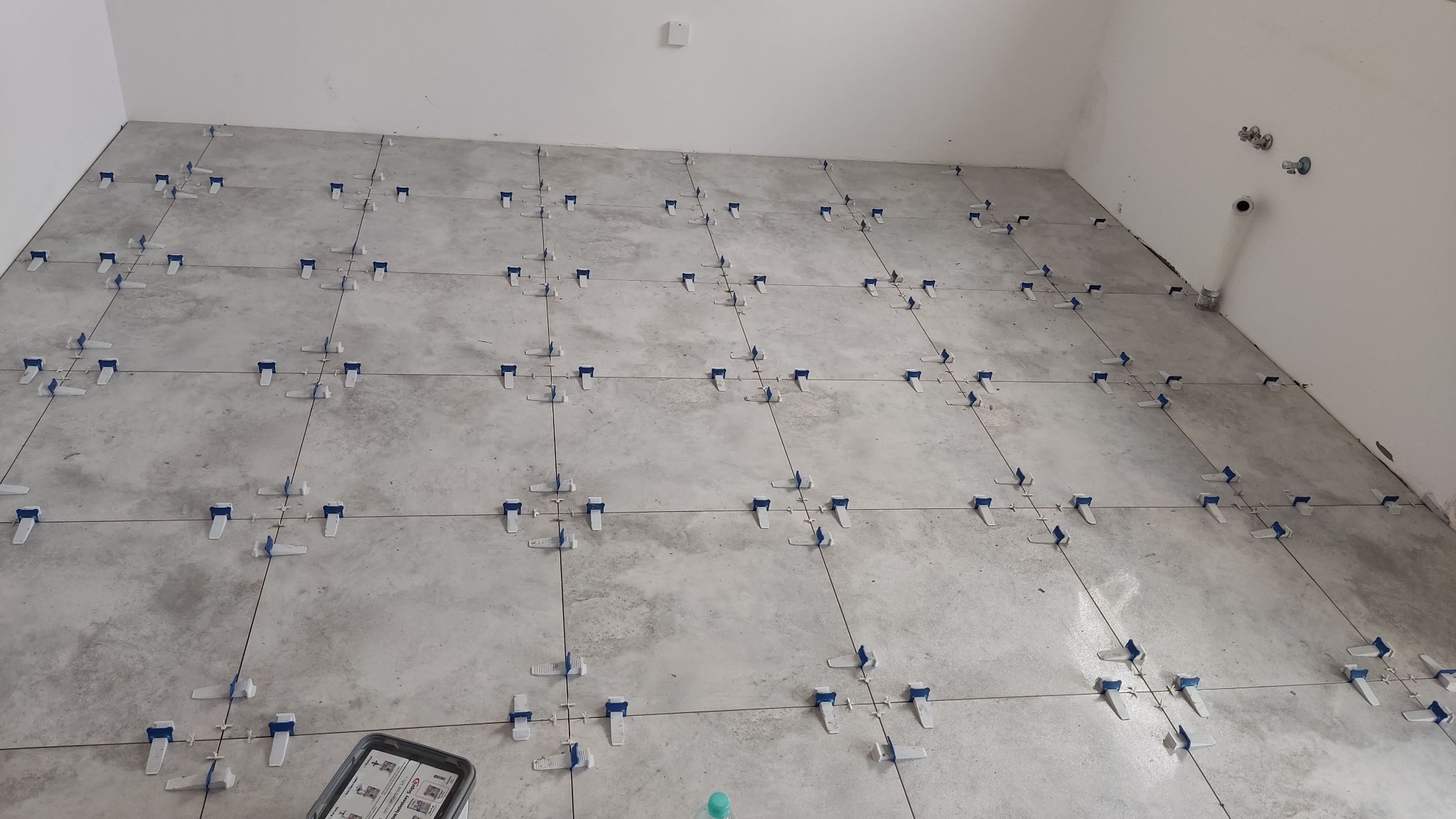 Szare płytki podłogowe w trakcie układania, z systemem poziomowania (klipsy i kliny). Widoczny fragment ściany i podłogi w pomieszczeniu.