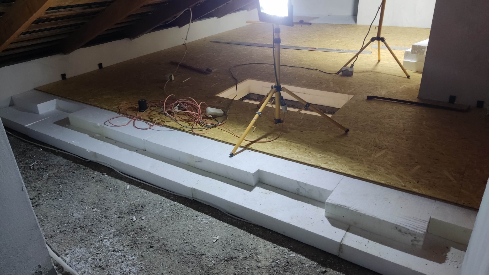 Ocieplenie poddasza: płyty OSB na legarach z białego styropianu, widoczne kable i lampa budowlana. Ukośny sufit z drewnianymi belkami.