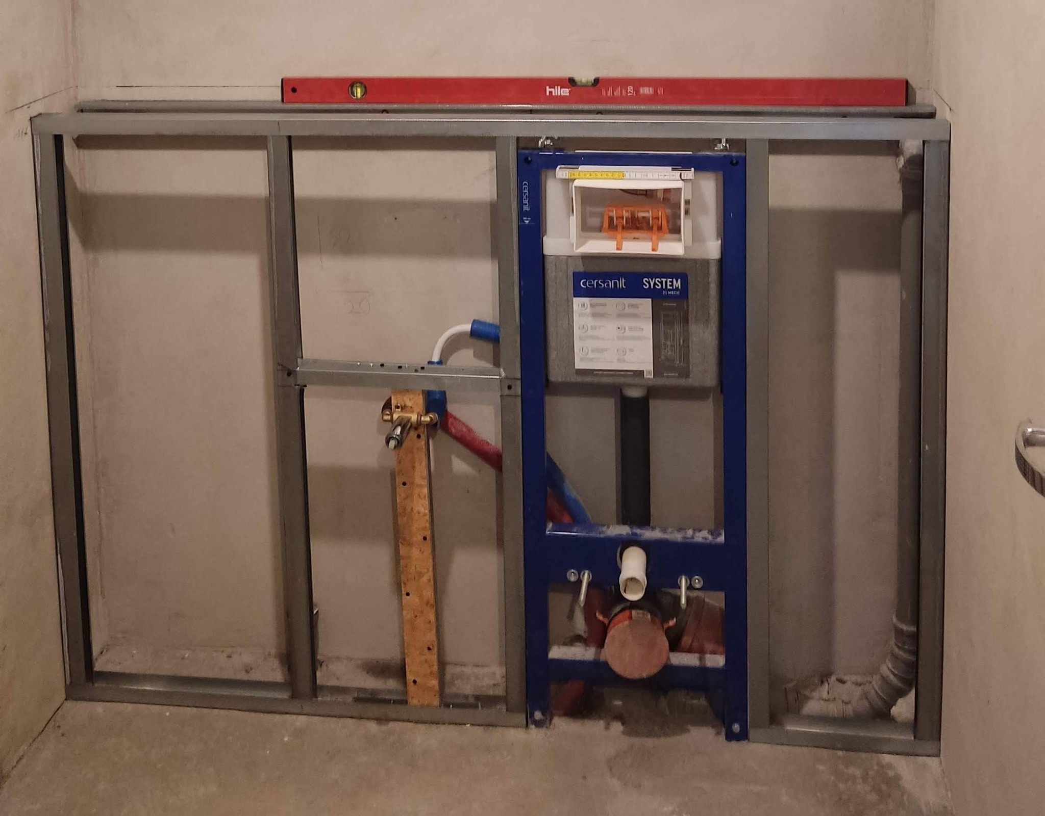 Stelaż podtynkowy Cersanit System zamontowany w metalowej konstrukcji, widoczne rury doprowadzające wodę. Poziomica Hiller na górnej belce. Etap prac w łazience.