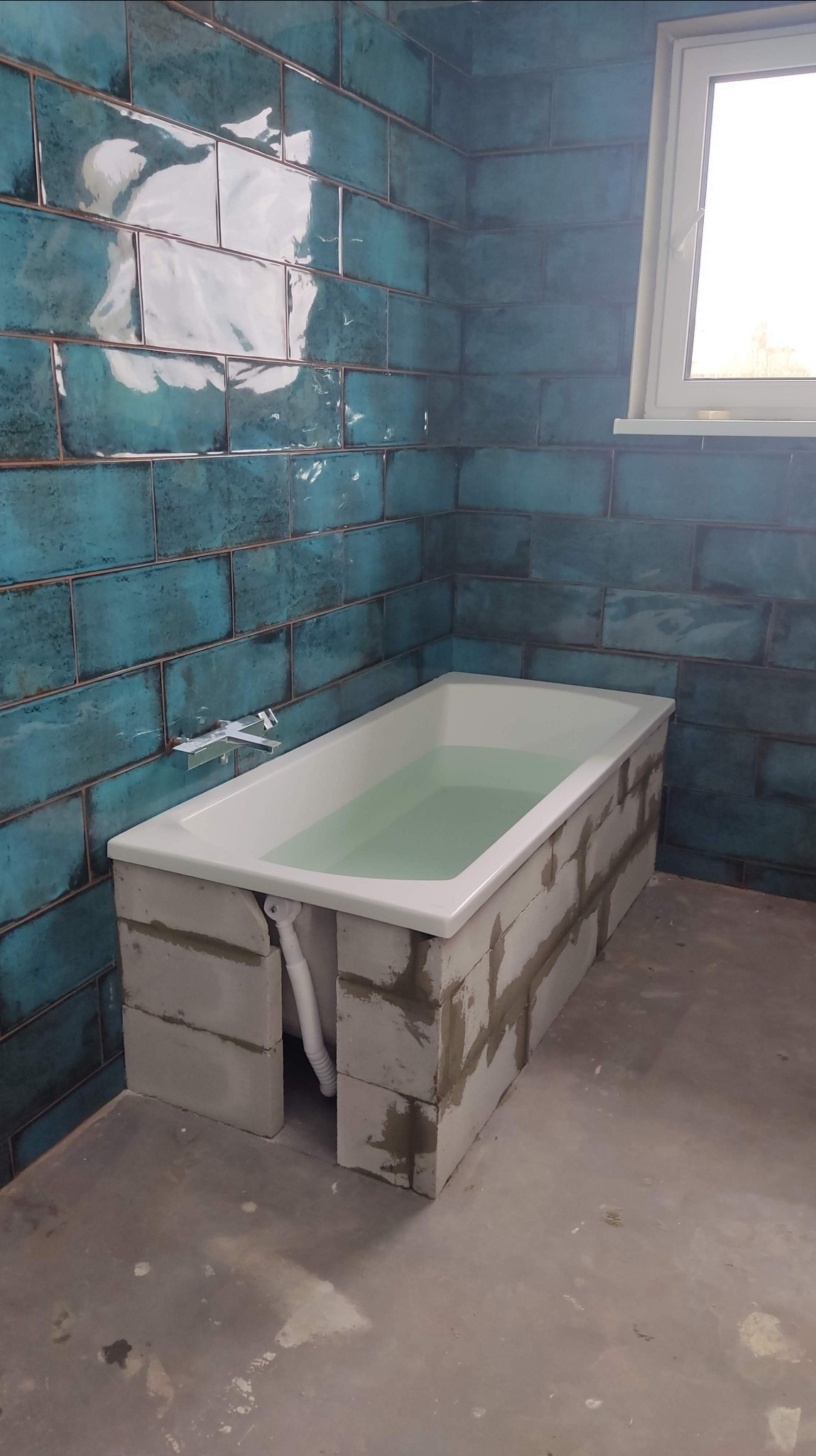 Niebieska, błyszcząca glazura na ścianie łazienki, obudowana wanna z białych bloczków, wypełniona wodą, widok na okno i surową podłogę.