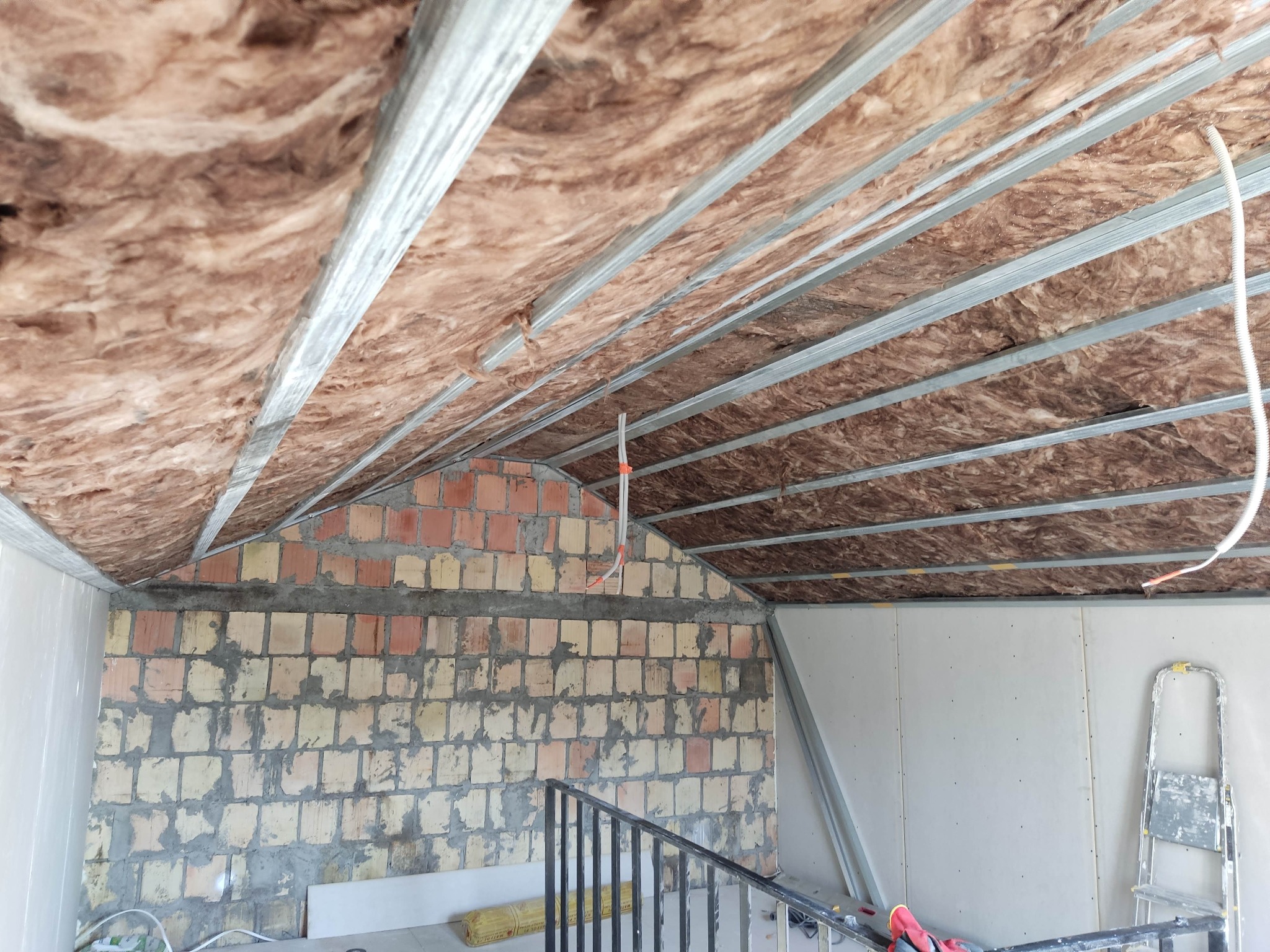 Skośny strych w trakcie izolacji: wełna mineralna między metalowymi profilami, surowa ściana z cegieł, częściowo obłożona płytami gipsowymi. Widoczna drabina.