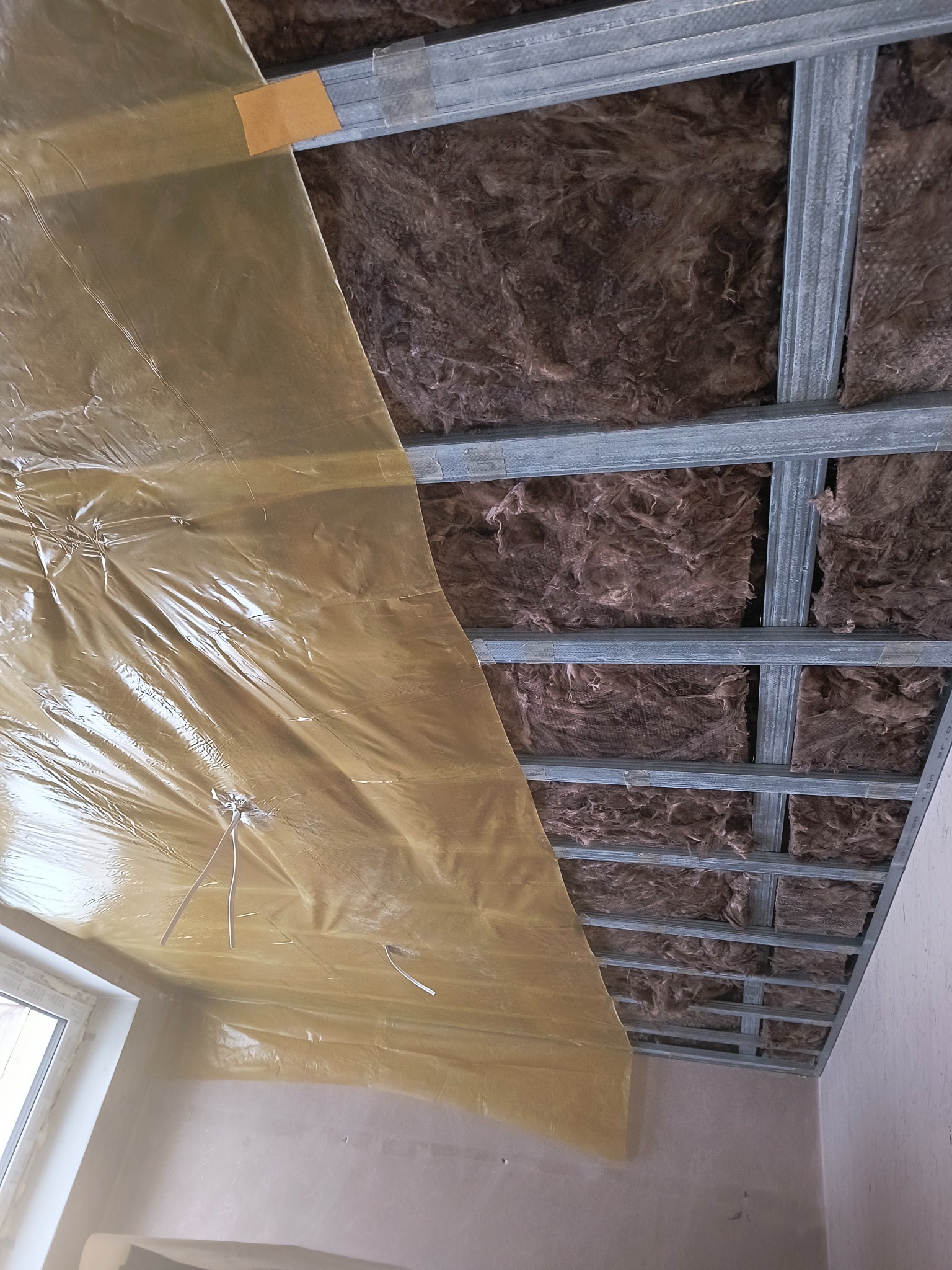 Montaż izolacji z wełny mineralnej i folii paroizolacyjnej na metalowym ruszcie sufitu podwieszanego w trakcie prac wykończeniowych wnętrza.