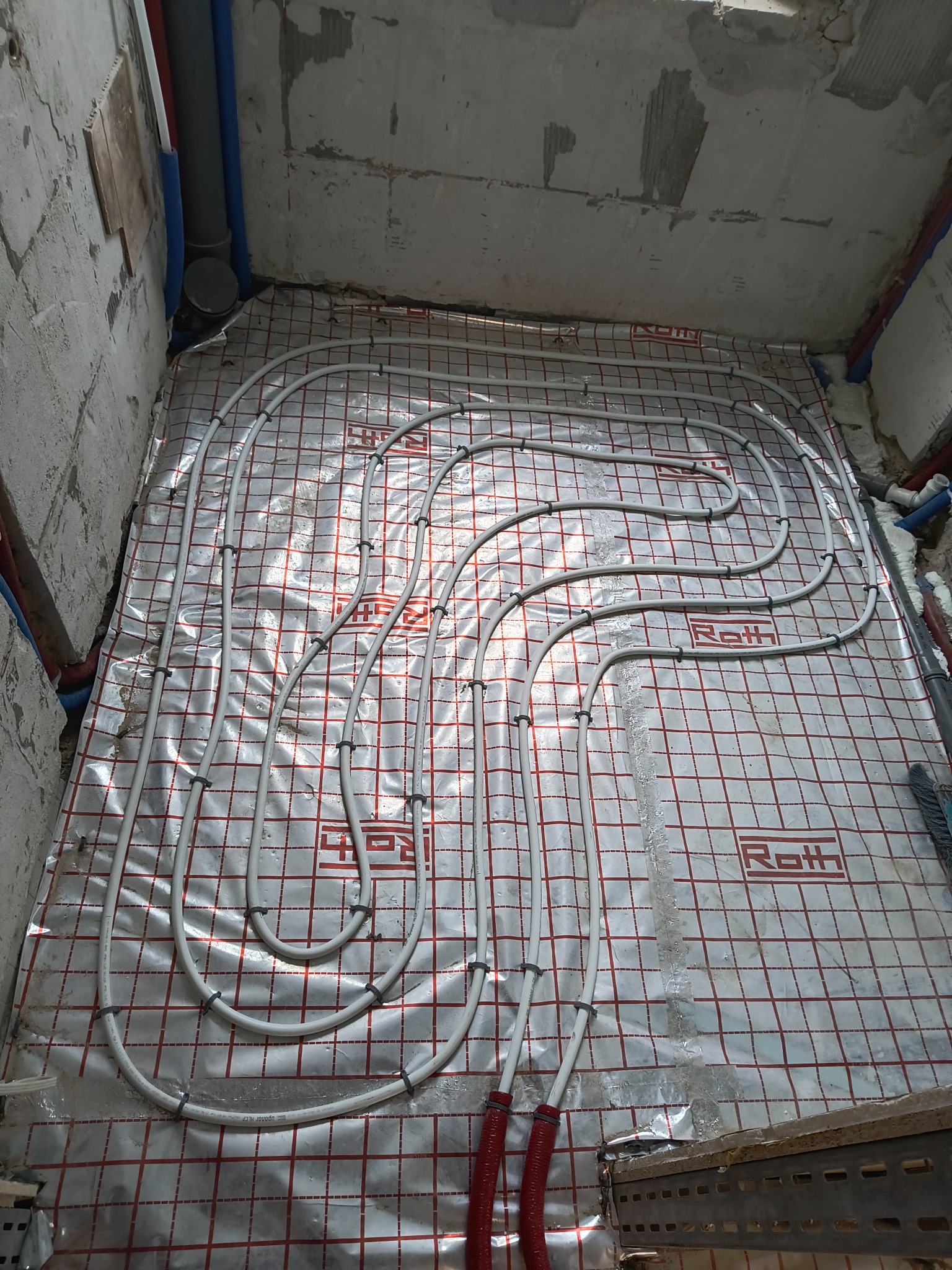 Ułożone rury ogrzewania podłogowego na folii izolacyjnej z siatką w surowym wnętrzu. Widoczne mocowania rur i fragmenty instalacji wodnej.