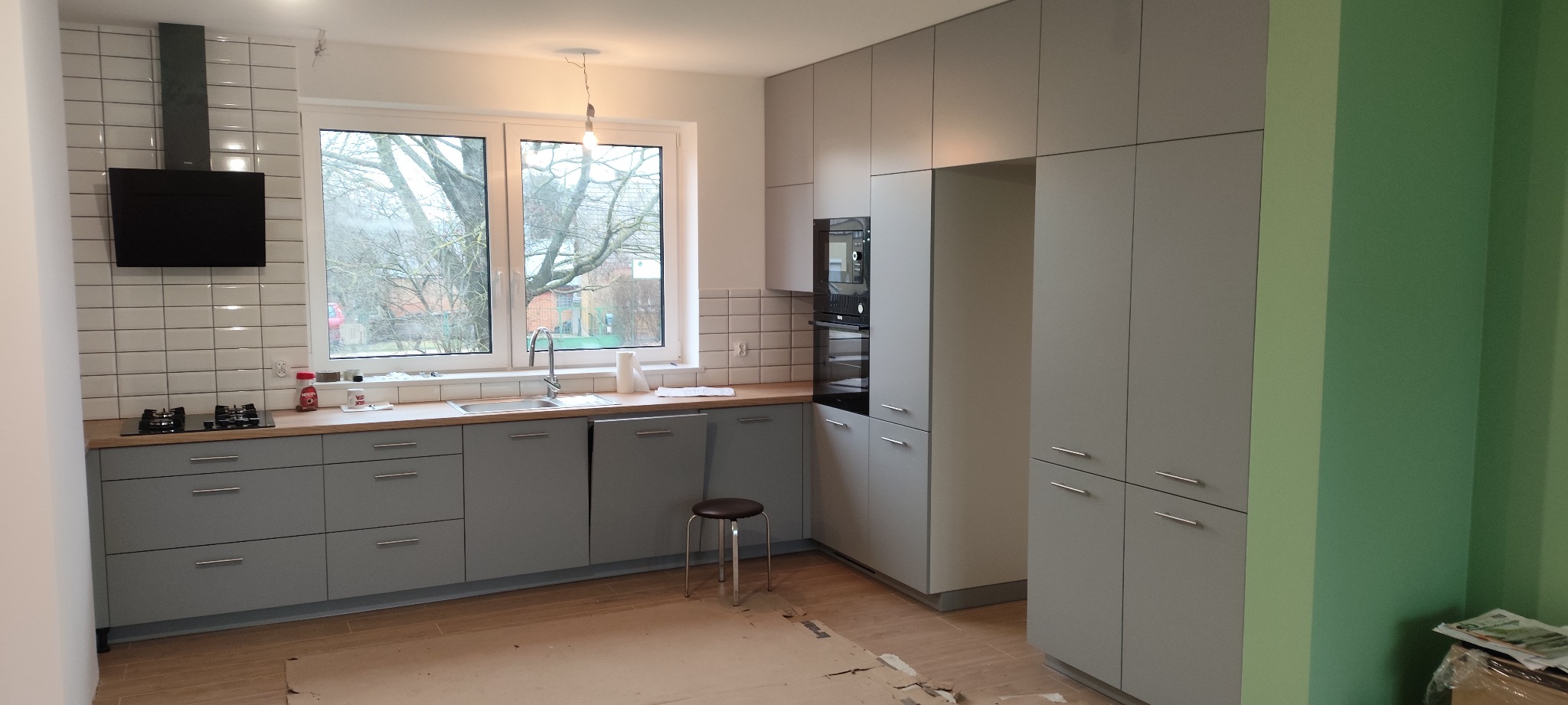 Nowoczesna kuchnia w szarościach z drewnianym blatem i białymi płytkami. Widoczny okap, okno z widokiem na drzewa i fragment zielonej ściany. Minimalistyczny design.