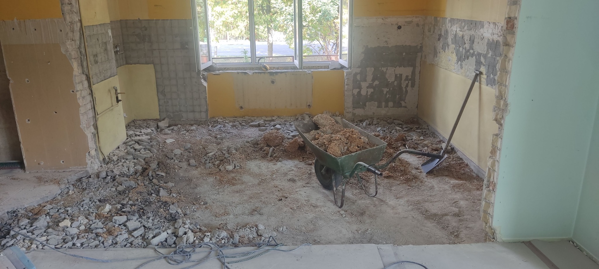 Demontaż płytek w kuchni: gruz na podłodze, taczka z resztkami, ściany częściowo skute, okno w tle. Etap przygotowawczy do remontu kuchni.