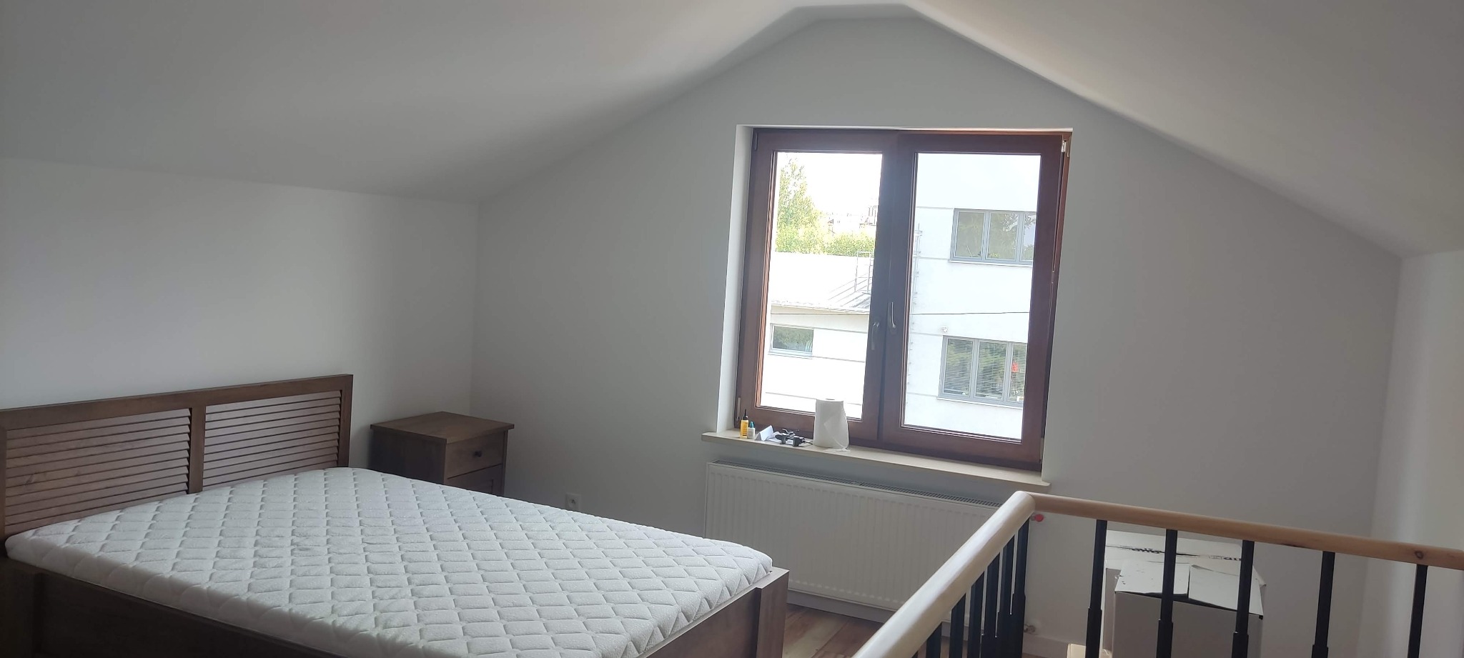 Jasne poddasze po remoncie: drewniane łóżko z materacem, szafka nocna, okno z widokiem na budynek. Świeżo pomalowane ściany i grzejnik. Widok z antresoli.