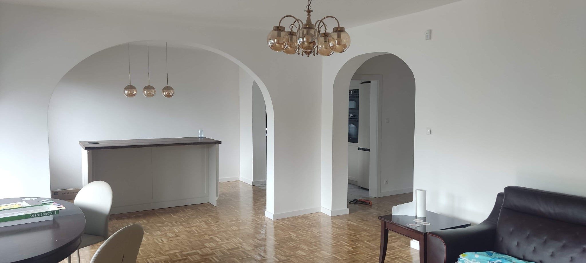 Jasne wnętrze z parkietem w jodełkę, łukowatymi przejściami i barkiem. Widoczny fragment sofy i stołu. Dwa rodzaje lamp wiszących. W tle widoczna kuchnia.