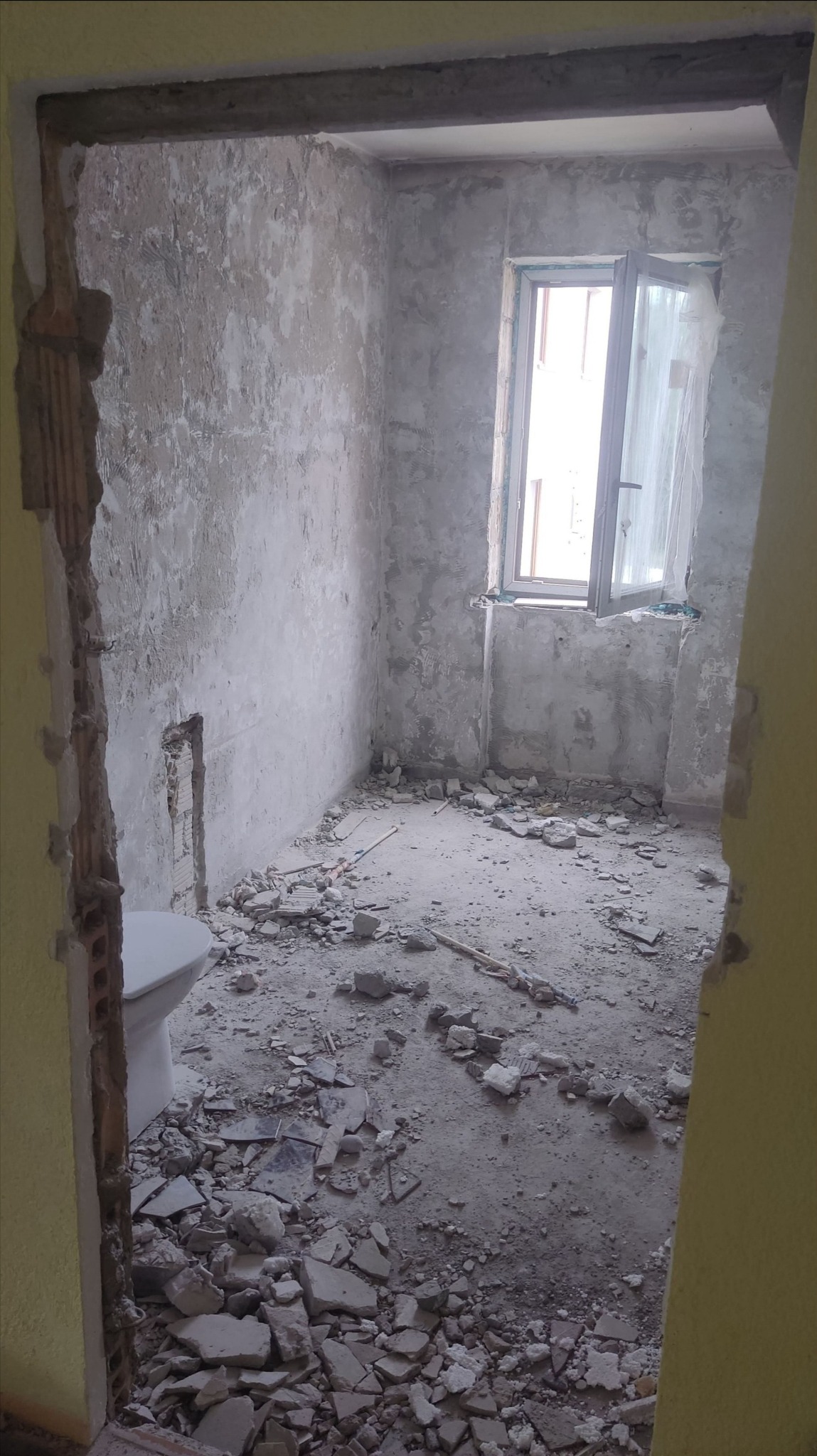 Łazienka w trakcie remontu: gruz na podłodze, ściany przygotowane do prac wykończeniowych, widoczne fragmenty instalacji. Okno z firanką w tle.