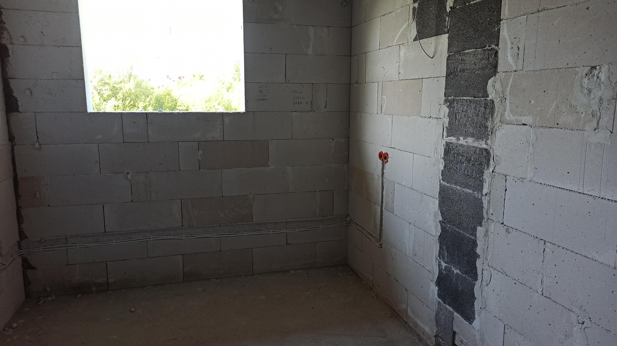 Surowy pokój z białych bloczków, widoczne okno, instalacja elektryczna i fragment ściany z szarych bloczków. Wnętrze w trakcie prac wykończeniowych.