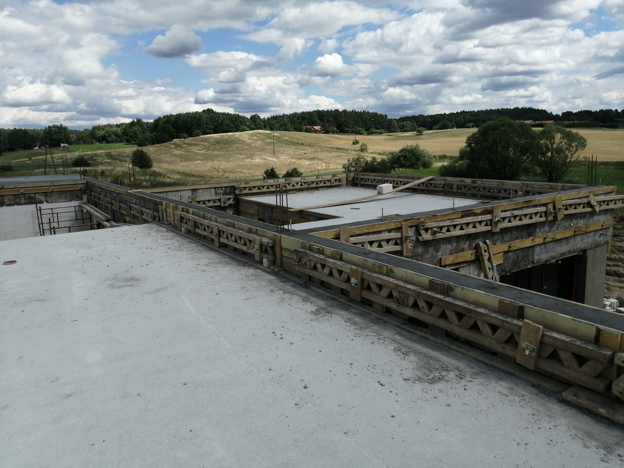 Zalany strop budynku w trakcie budowy, widok z góry na drewniane szalunki i beton, w tle krajobraz z polami i lasem pod błękitnym niebem.