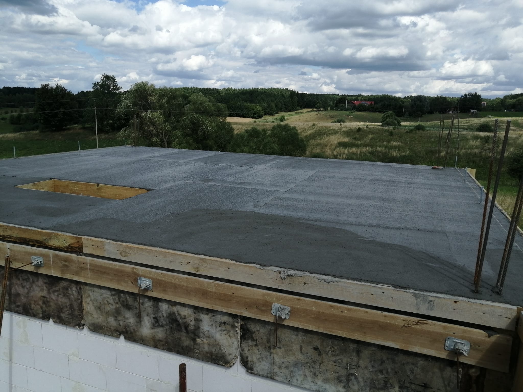 Wylany strop betonowy z otworem, widoczne zbrojenia i szalunki. W tle zielony krajobraz z lasem i pochmurnym niebem. Budowa domu jednorodzinnego.