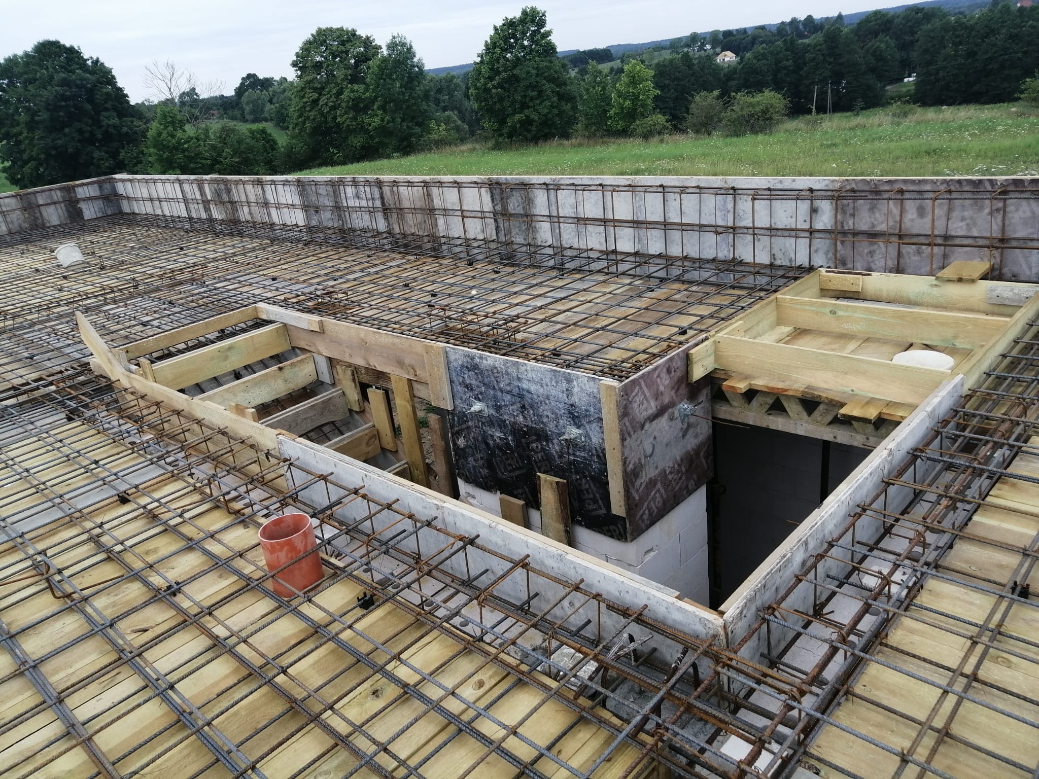 Zbrojenie stropu budynku w trakcie budowy, widoczne drewniane szalunki, stalowe pręty i fragment krajobrazu w tle. Pomarańczowy element instalacji na pierwszym planie.