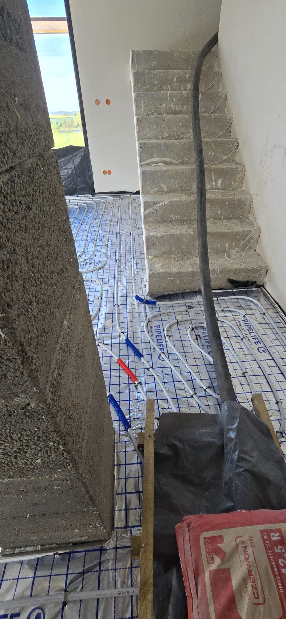 Montaż ogrzewania podłogowego w Ługwałdzie: rury grzewcze ułożone na folii izolacyjnej, widoczne elementy instalacji i schody w surowym stanie.