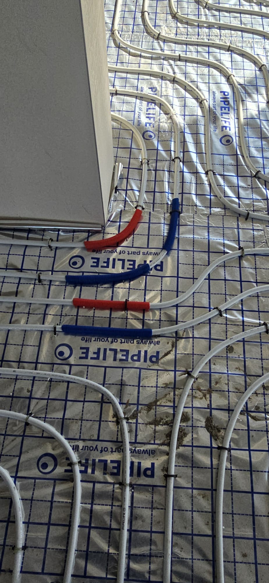 Białe rurki ogrzewania podłogowego, częściowo w izolacji czerwonej i niebieskiej, ułożone na folii z nadrukiem, widok z góry, etap instalacji.