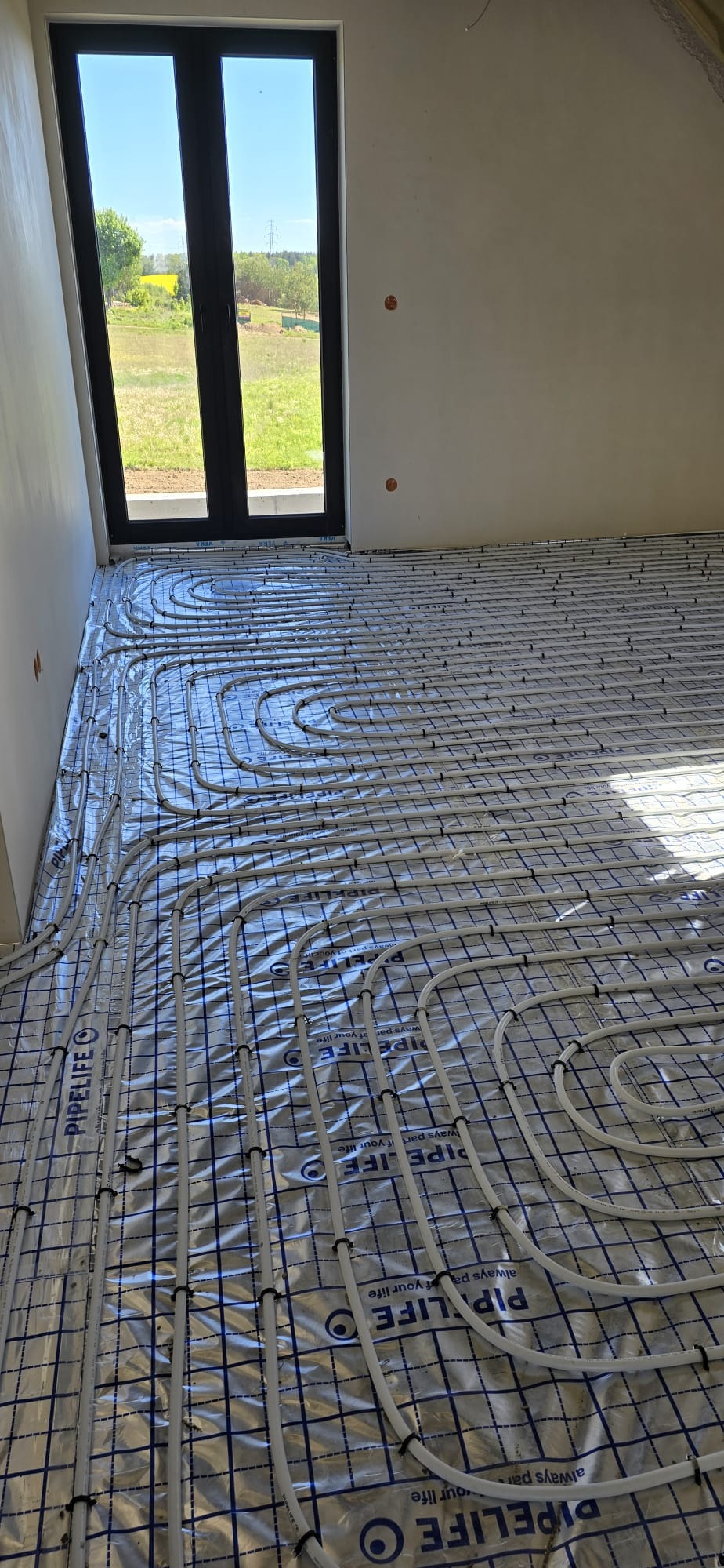 Białe rury ogrzewania podłogowego ułożone na folii z nadrukiem PIPELIFE, widok z góry, w tle okno z widokiem na zewnątrz, surowy stan wykończenia wnętrza.