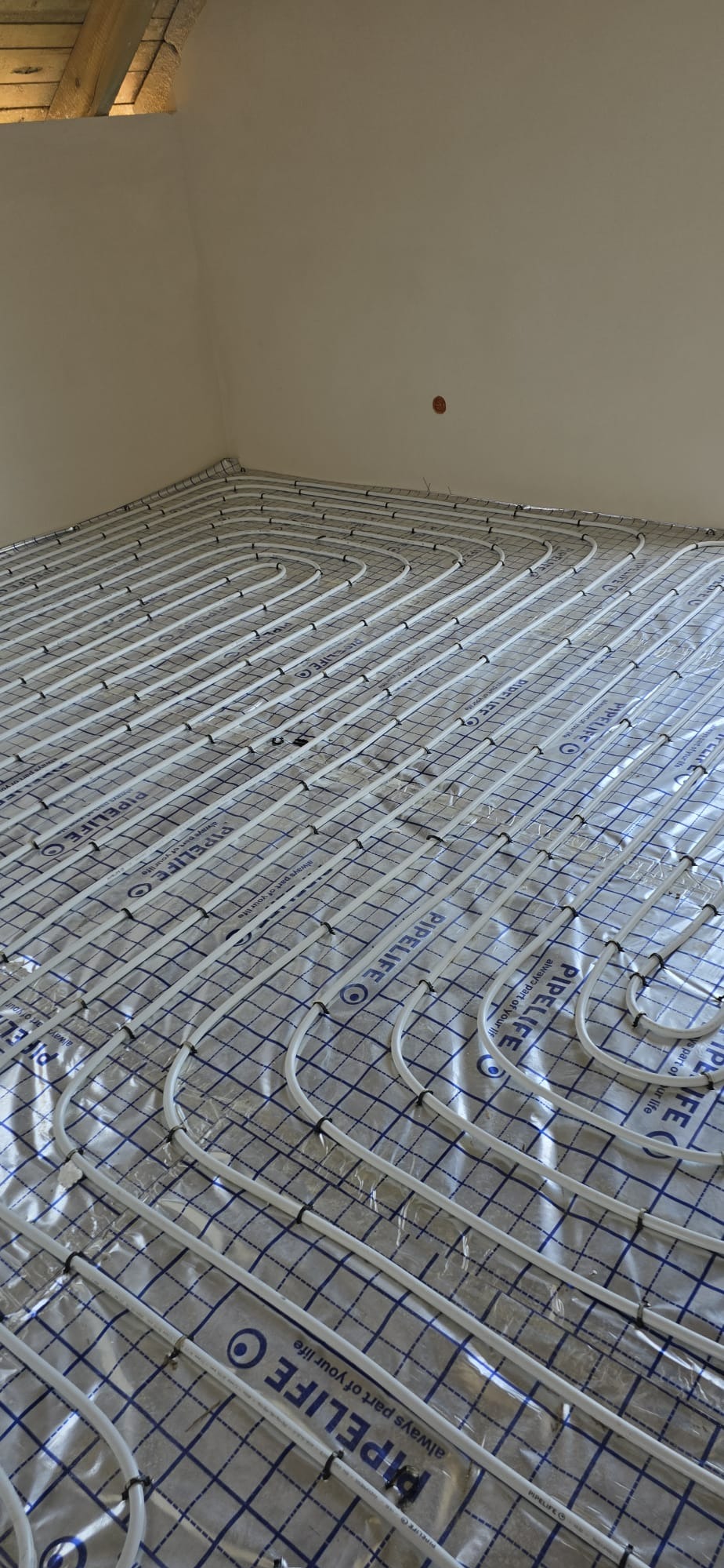 Białe rury ogrzewania podłogowego ułożone na folii z niebieską kratką, widok z góry, gotowe do zalania posadzką. Równoległe ułożenie rur.
