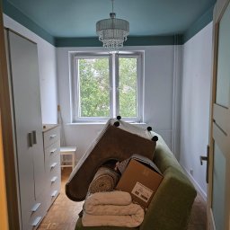 Pokój przed remontem: meble i rzeczy osobiste zgromadzone na kanapie, widok na okno, sufit pomalowany na niebiesko. Przygotowania do prac remontowych.