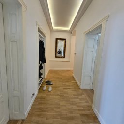 Korytarz z drewnianą podłogą i białymi ścianami. Po lewej stronie szafa z ubraniami, na końcu lustro w złotej ramie, w którym odbija się osoba. Oświetlenie sufitowe.