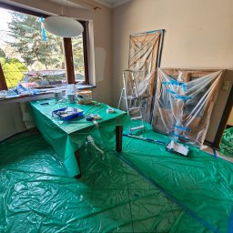 Pokój przygotowany do malowania: podłoga i drzwi zabezpieczone folią, stół z farbami i narzędziami malarskimi. Widoczna drabina i częściowo pomalowana ściana.