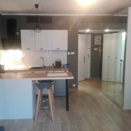 Nowoczesna kuchnia z wyspą i hokerem, białe szafki, szare ściany, drewnopodobna podłoga, lustrzana szafa w tle. Minimalistyczny design, oświetlenie punktowe.