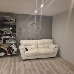 SUNSHINE SPÓŁKA Z OGRANICZONĄ ODPOWIEDZIALNOŚCIĄ - Szara ściana z efektem betonu w pokoju w Poznaniu, obok biała sofa i regał z ubraniami. Podłoga z paneli. Minimalistyczne wnętrze.