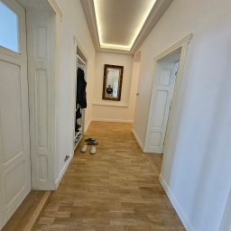 SUNSHINE SPÓŁKA Z OGRANICZONĄ ODPOWIEDZIALNOŚCIĄ - Jasny korytarz z parkietem i białymi ścianami. Widoczne drzwi z ozdobnymi framugami, lustro w złotej ramie i oświetlenie sufitowe. W tle szafa z ubraniami.