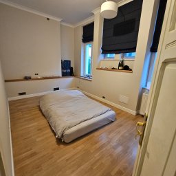 SUNSHINE SPÓŁKA Z OGRANICZONĄ ODPOWIEDZIALNOŚCIĄ - Pokój z drewnianą podłogą i łóżkiem. Widoczne okna z roletami rzymskimi, półka pod oknem z butelką i szklankami. Ściany w jasnym odcieniu beżu.