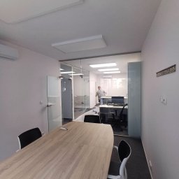 SUNSHINE SPÓŁKA Z OGRANICZONĄ ODPOWIEDZIALNOŚCIĄ - Jasne biuro po remoncie: szklane ściany działowe, nowoczesne oświetlenie, stół konferencyjny. W tle pracownik sprzątający, co sugeruje świeżo zakończone prace wykończeniowe.