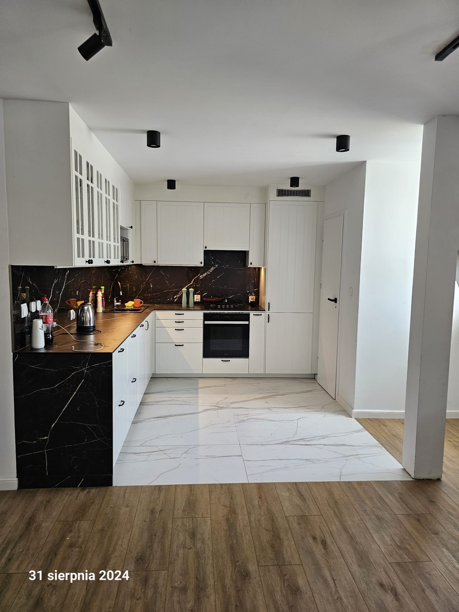 Nowoczesna kuchnia z białymi szafkami i blatem z czarnego marmuru. Podłoga wyłożona białymi płytkami z szarymi żyłami. Czarne lampy sufitowe. Widok na drewnianą podłogę w sąsiednim pomieszczeniu.