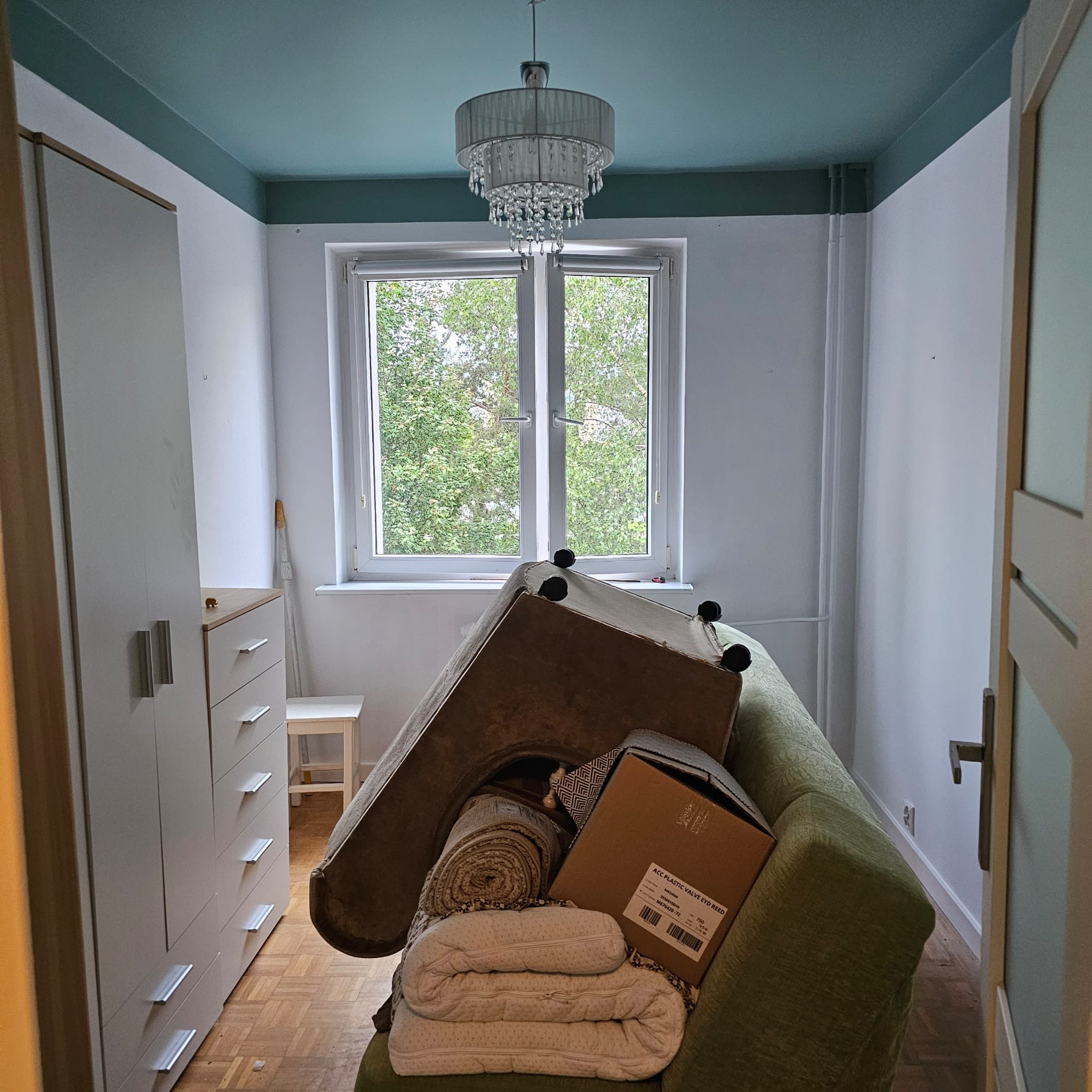 Pokój przed remontem: meble i rzeczy osobiste zgromadzone na kanapie, widok na okno, sufit pomalowany na niebiesko. Przygotowania do prac remontowych.