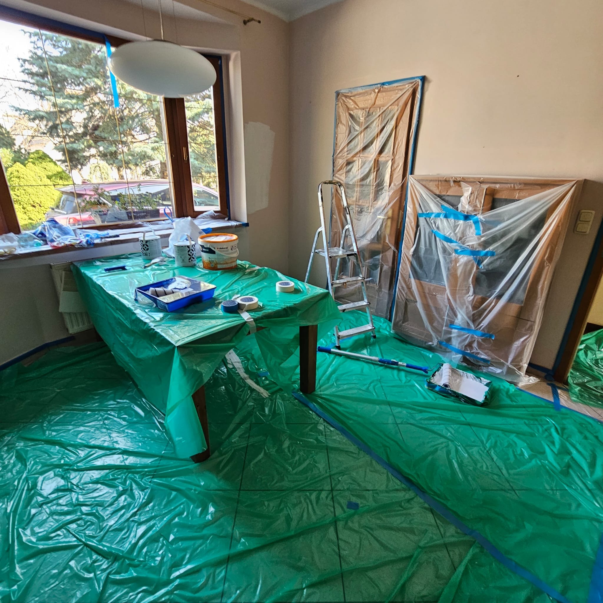 Pokój przygotowany do malowania: podłoga i drzwi zabezpieczone folią, stół z farbami i narzędziami malarskimi. Widoczna drabina i częściowo pomalowana ściana.