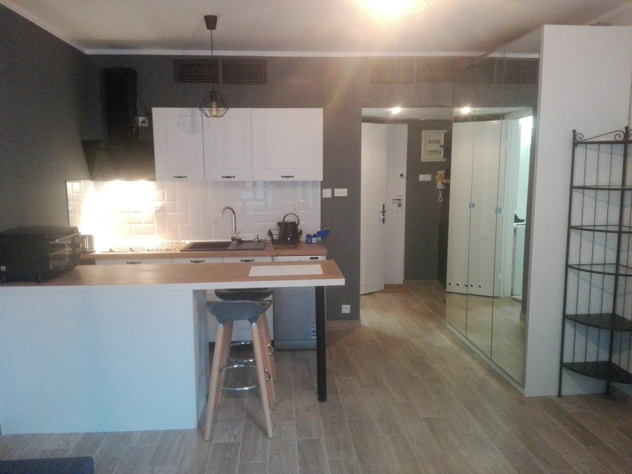 Nowoczesna kuchnia z wyspą i hokerem, białe szafki, szare ściany, drewnopodobna podłoga, lustrzana szafa w tle. Minimalistyczny design, oświetlenie punktowe.