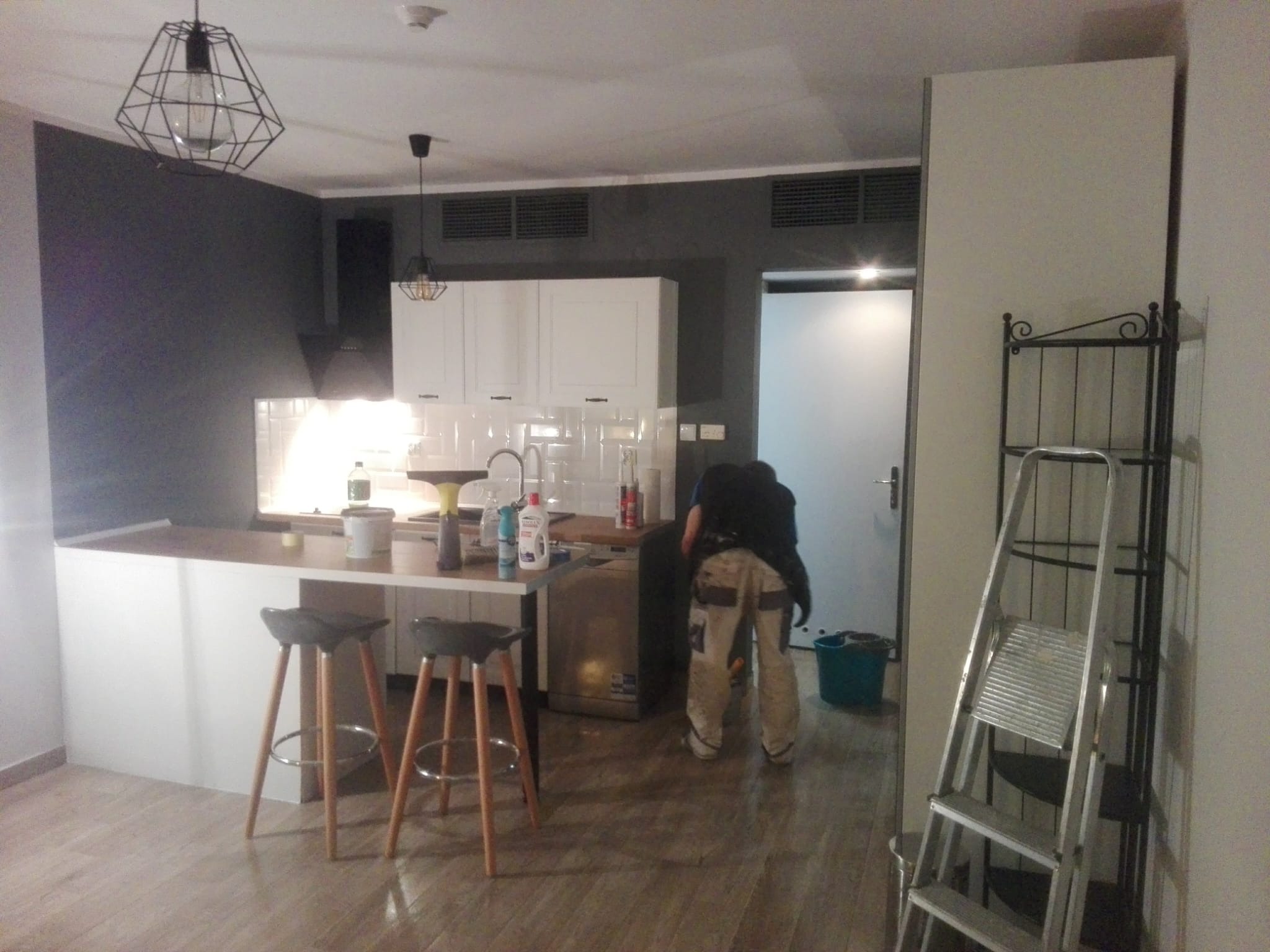 Wnętrze kuchni w trakcie prac wykończeniowych. Widoczny pracownik w trakcie pracy, drabina, farby i akcesoria malarskie. Nowoczesne oświetlenie i wykończenie.