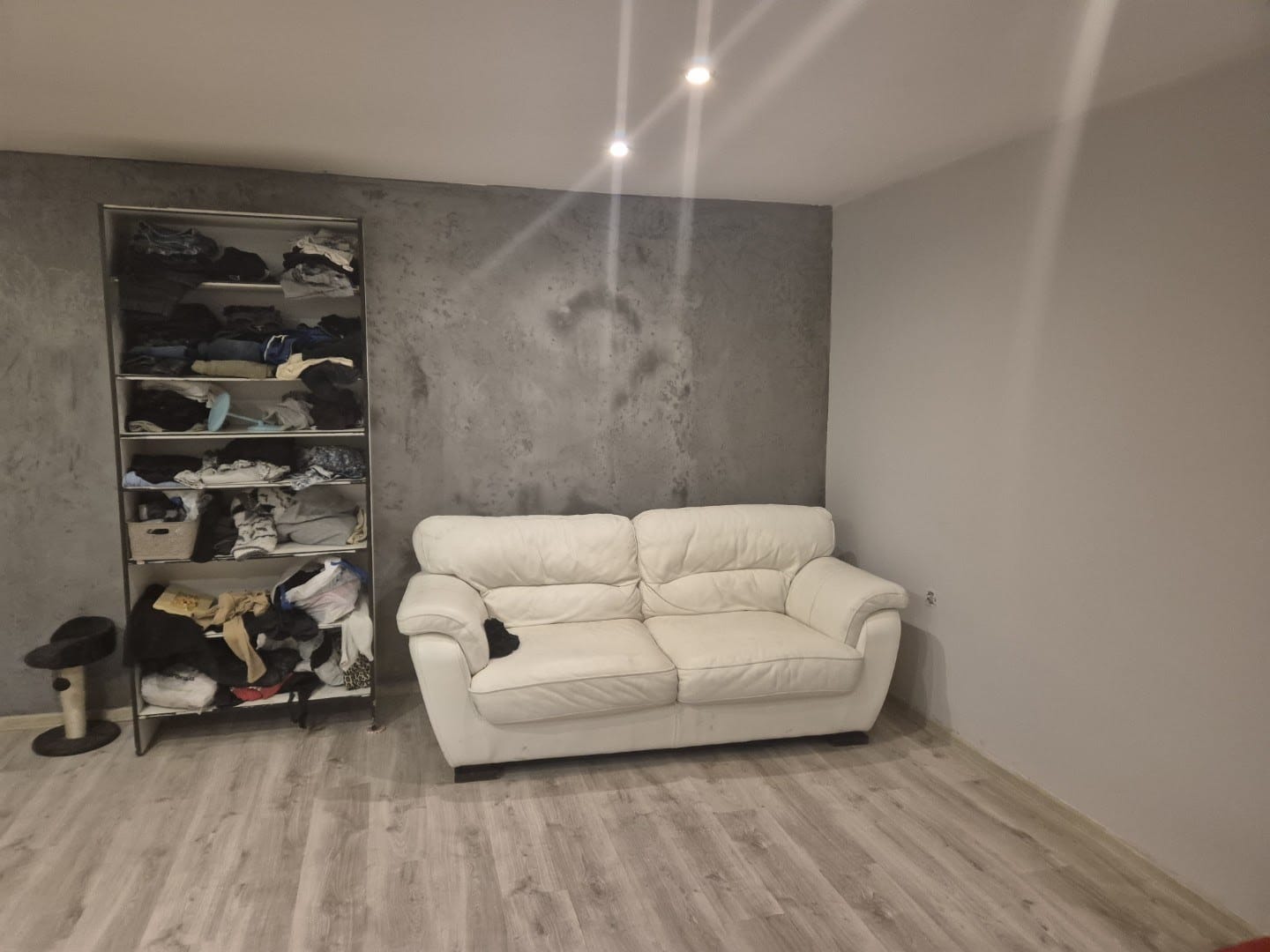 Szara ściana z efektem betonu w pokoju w Poznaniu, obok biała sofa i regał z ubraniami. Podłoga z paneli. Minimalistyczne wnętrze.