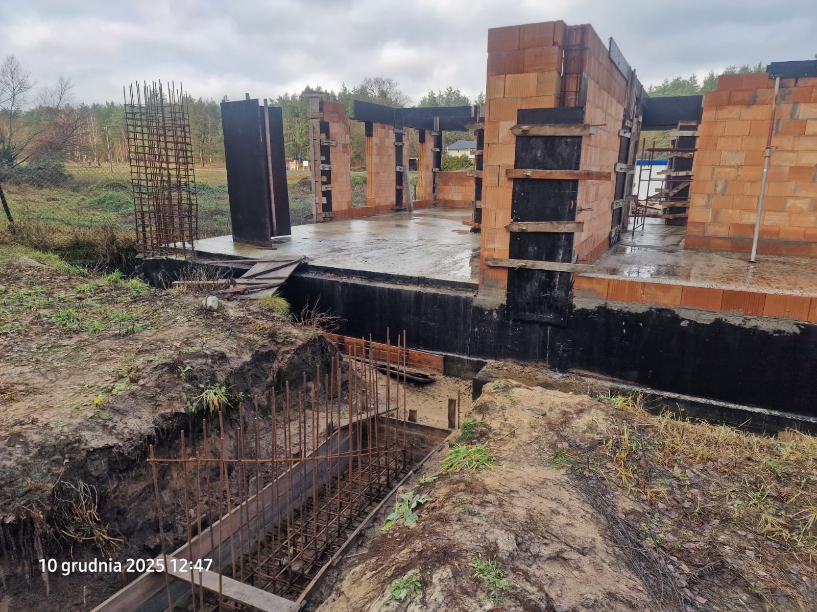 Budowa domu w trakcie: fundamenty z izolacją, ściany z pustaków ceramicznych, zbrojenie w wykopie. Widok na plac budowy w pochmurny dzień.