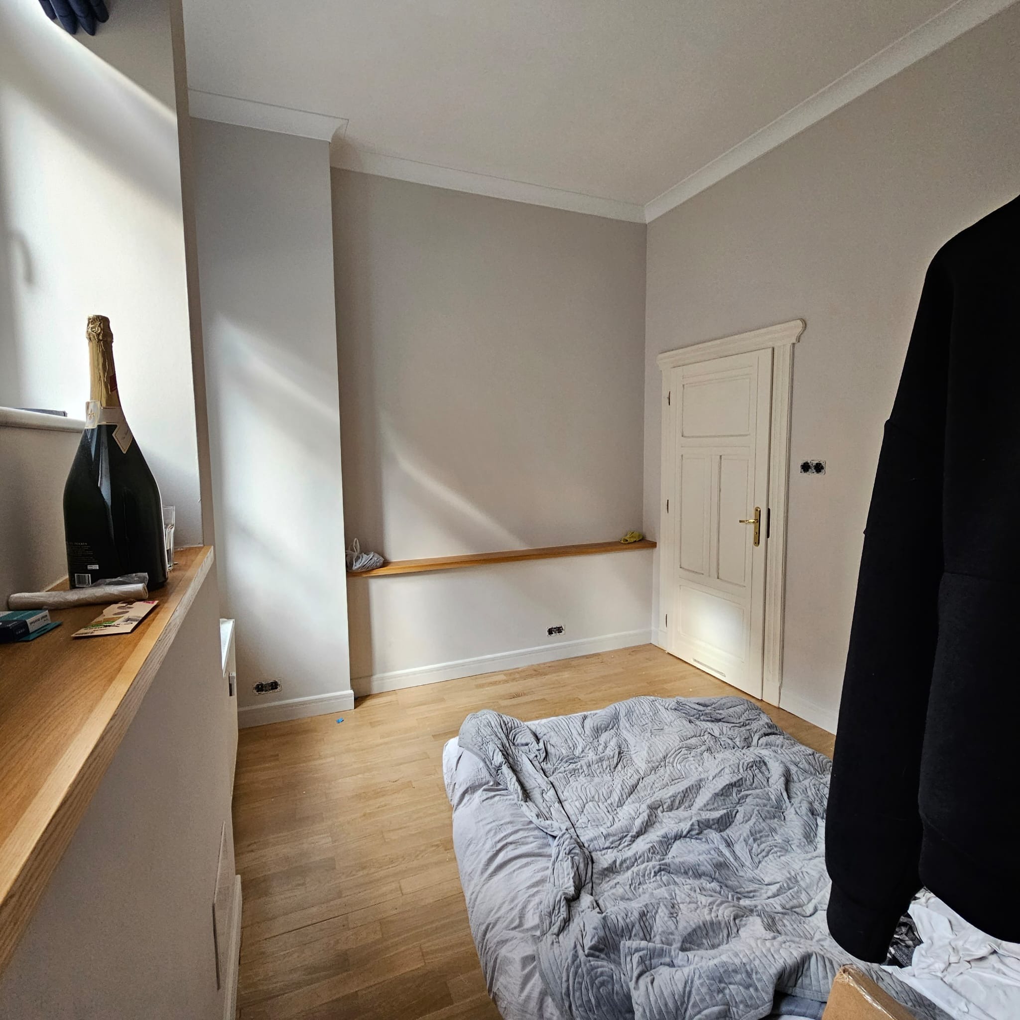 Pokój przed remontem: drewniana podłoga, szare ściany, białe listwy przypodłogowe i drzwi. Na parapecie butelka szampana. Widoczne gniazdka elektryczne.