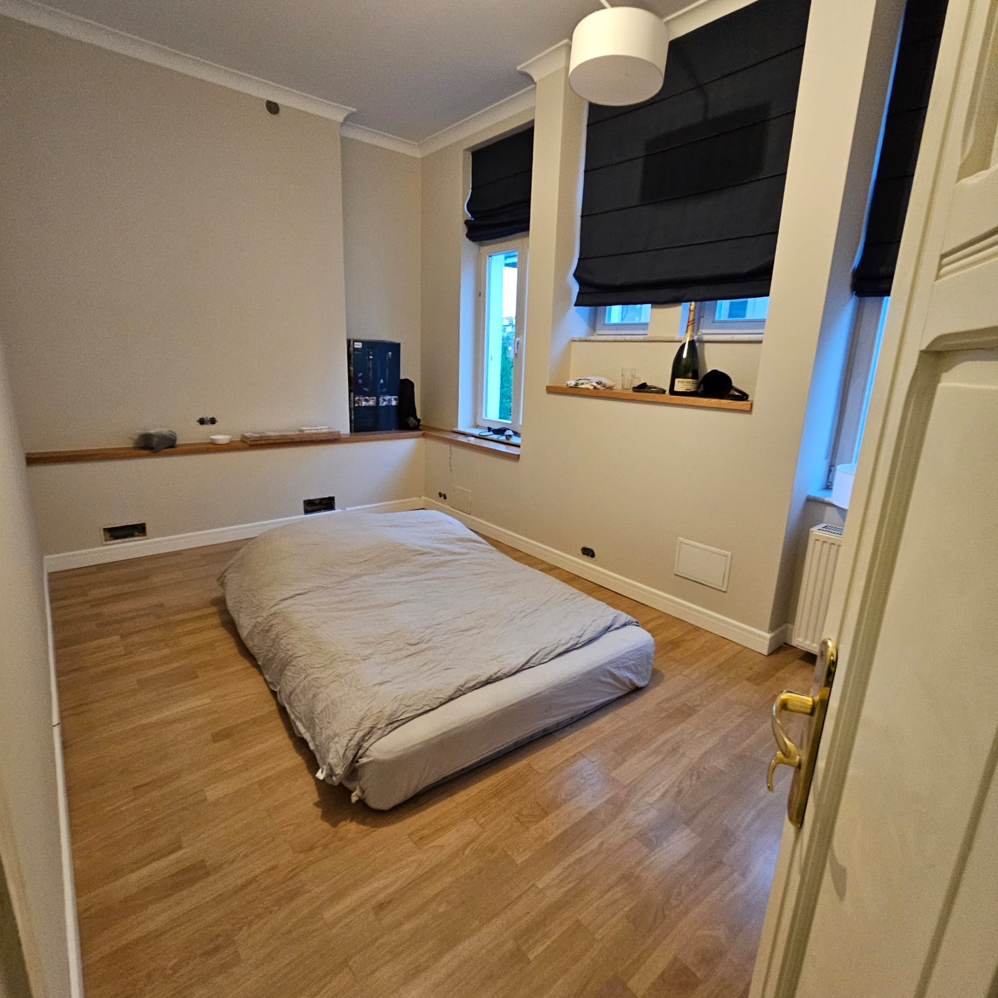 Pokój z drewnianą podłogą i łóżkiem. Widoczne okna z roletami rzymskimi, półka pod oknem z butelką i szklankami. Ściany w jasnym odcieniu beżu.