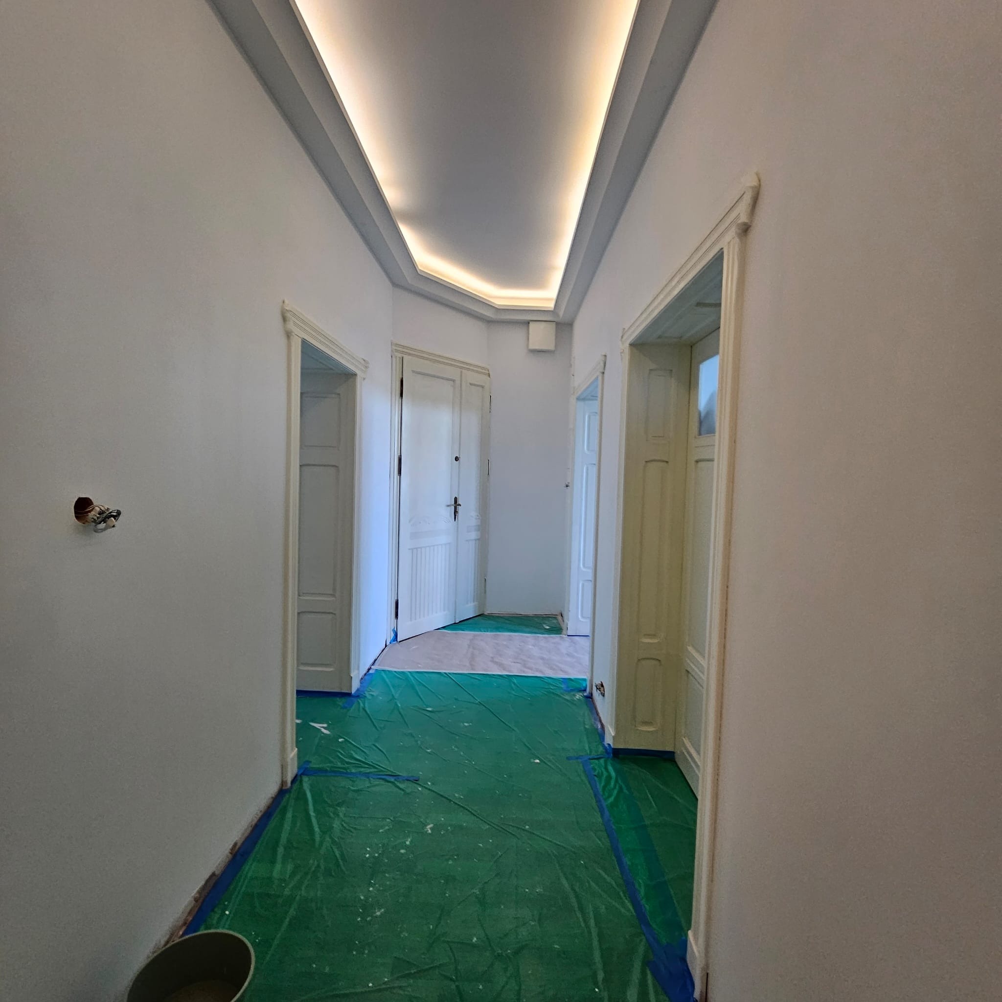 Białe, odświeżone wnętrze korytarza z trzema parami drzwi, zabezpieczone folią malarską. Sufit z podświetleniem LED. Gładkie ściany po malowaniu.