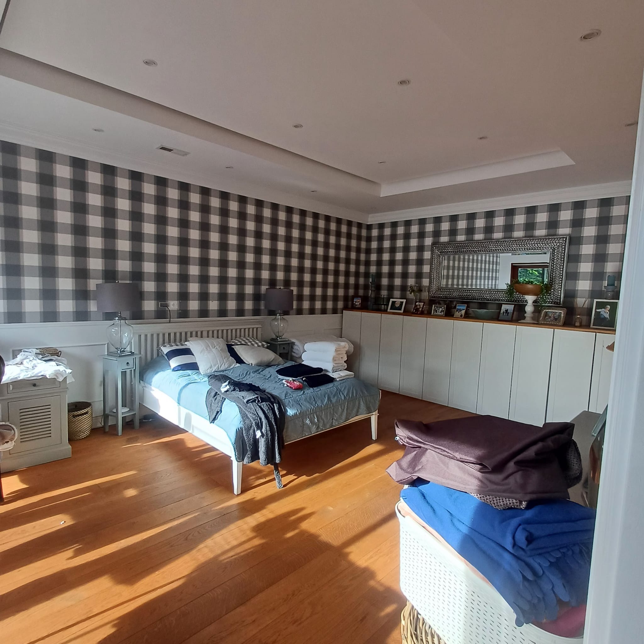 Sypialnia po remoncie: ściany w szaro-białą kratę, drewniana podłoga, białe meble. Widoczne łóżko i komoda. Duże okno wpuszcza promienie słoneczne.