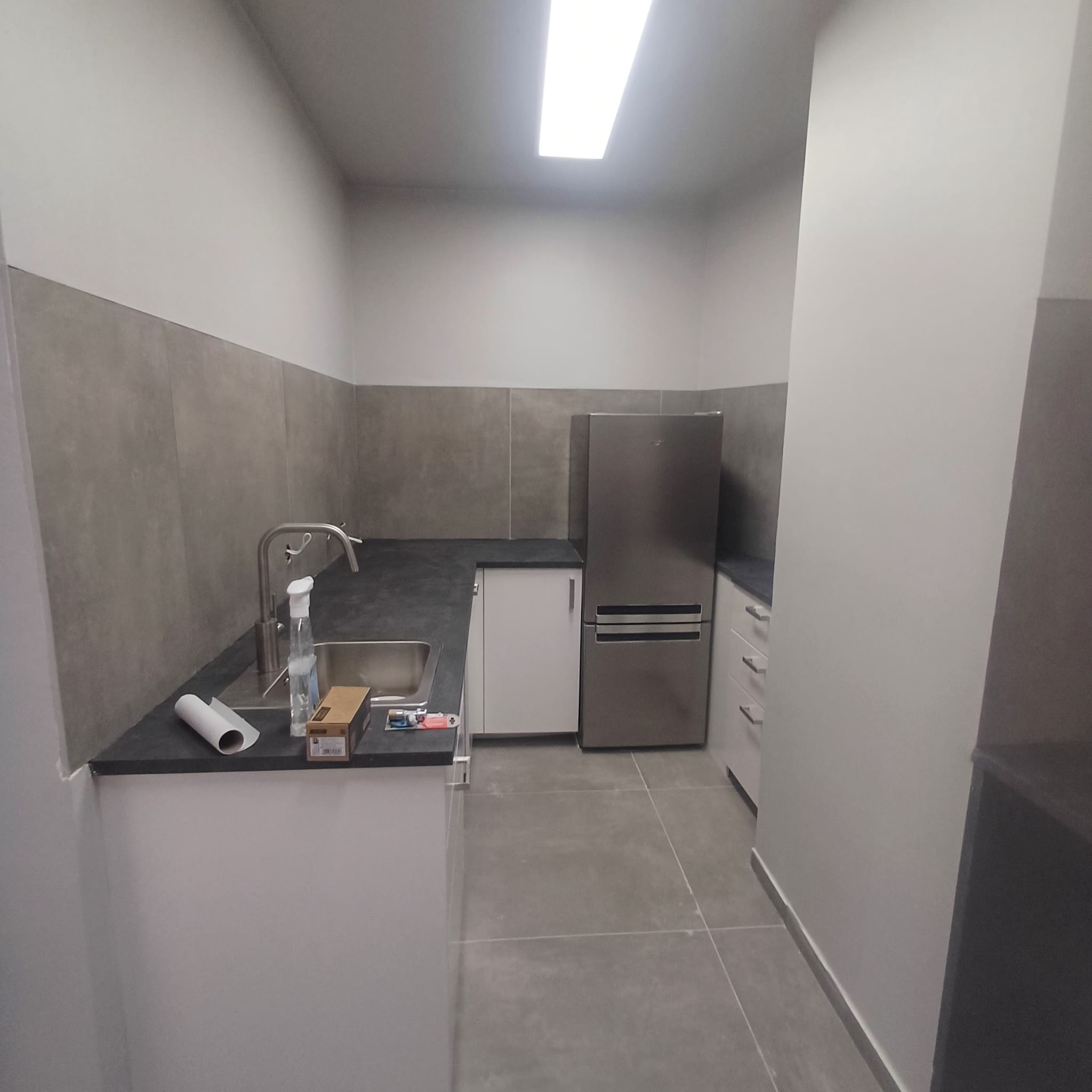 Nowoczesna kuchnia po remoncie: szare płytki na ścianie, białe szafki, srebrna lodówka. Minimalistyczny design, czysty blat z akcesoriami. Oświetlenie sufitowe.