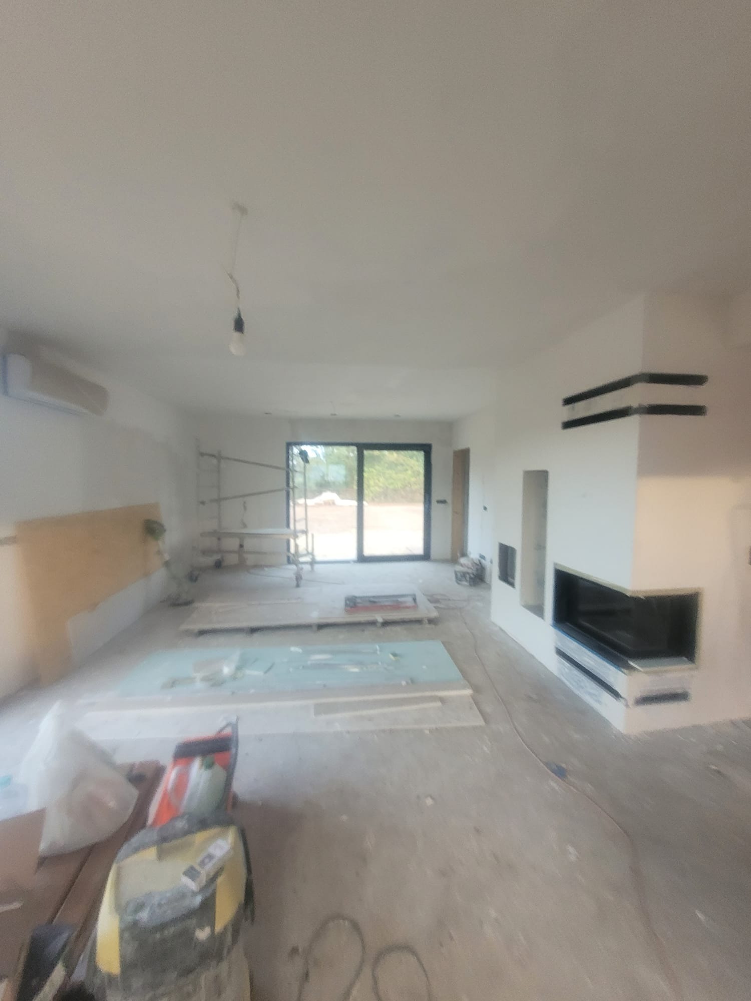 Wnętrze w trakcie prac wykończeniowych: widoczne rusztowanie, płyty gipsowe na podłodze i obudowa kominka z czarnymi akcentami. Surowy stan, przygotowanie do dalszych prac.