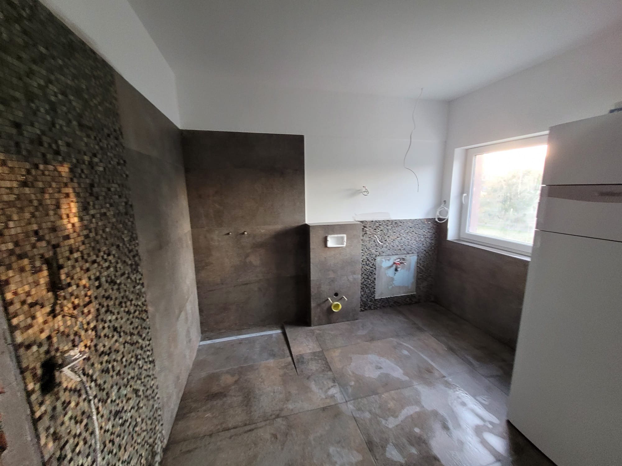 Wnętrze łazienki w trakcie prac wykończeniowych. Widoczne brązowe płytki na podłodze i ścianach, mozaika przy oknie oraz instalacje. Częściowo wykończone.