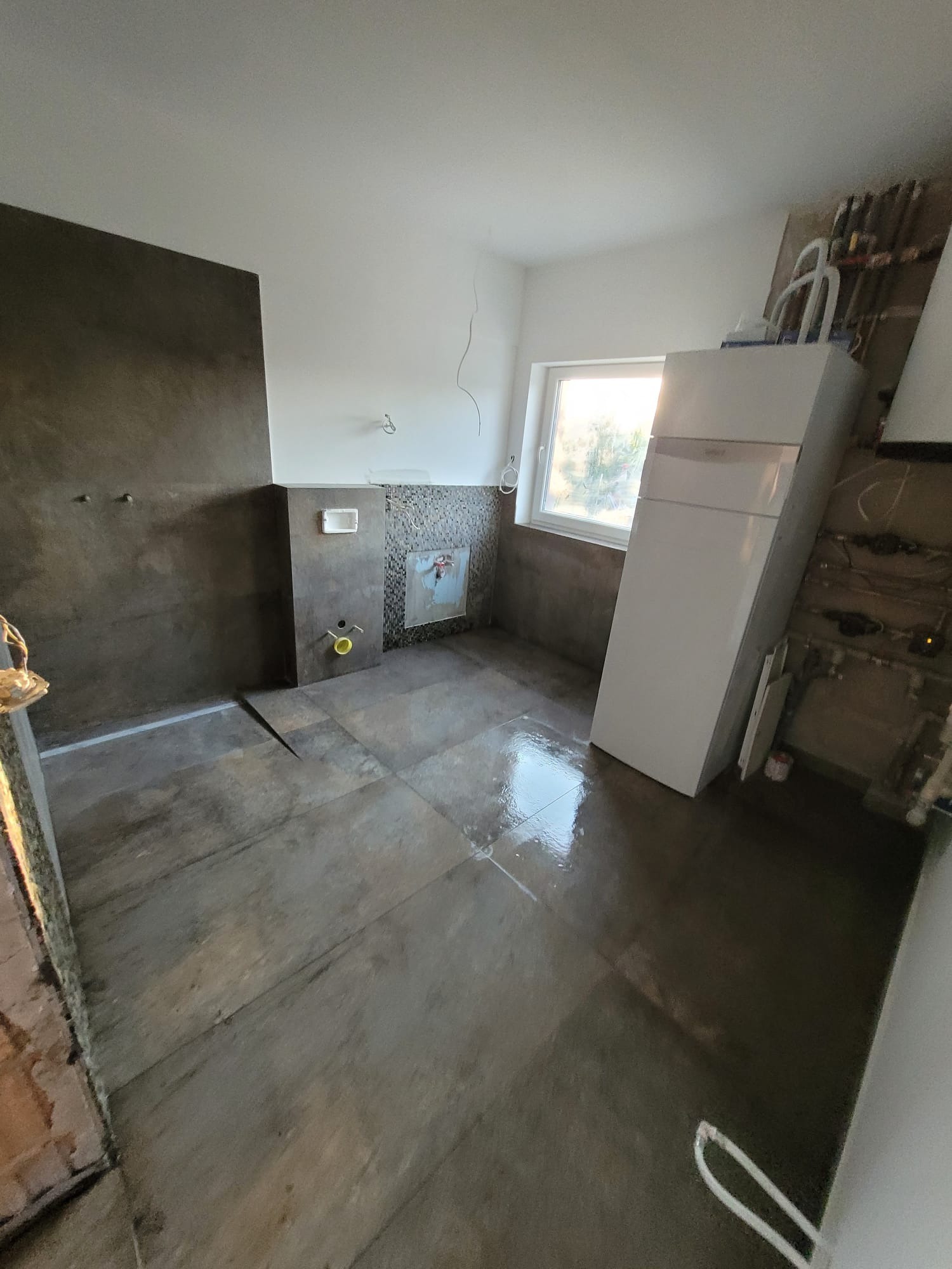 Wnętrze łazienki w trakcie prac wykończeniowych: położona podłoga z płytek imitujących drewno, częściowo wykończone ściany, widoczny bojler i instalacja.