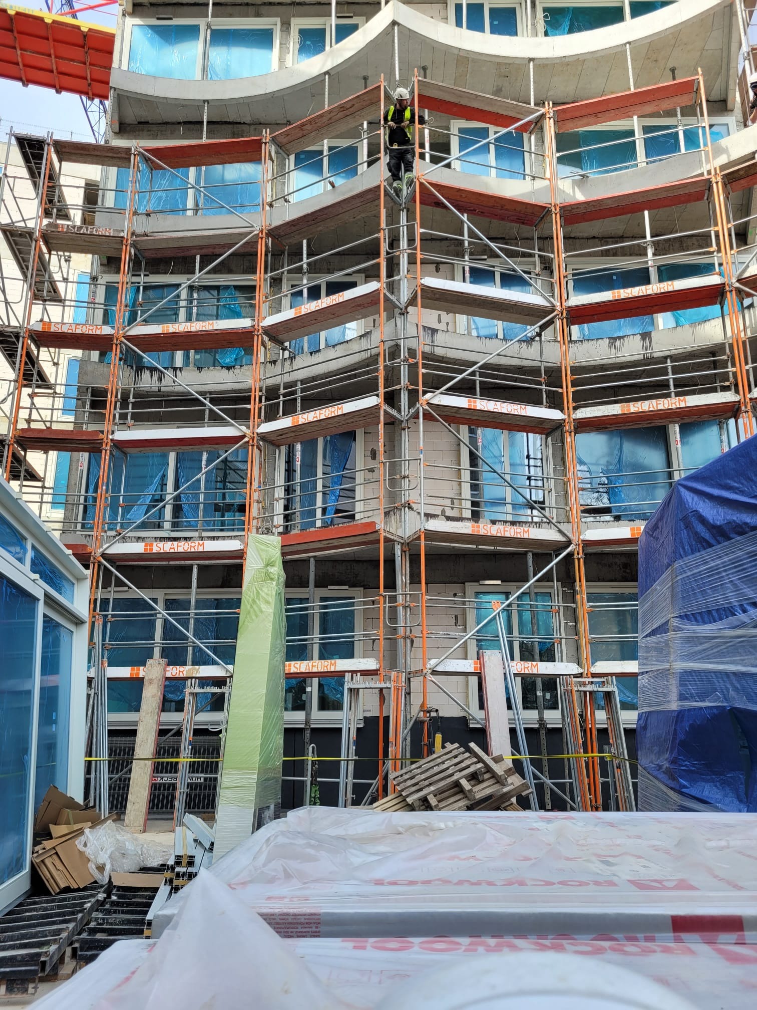 Budynek w trakcie budowy z rusztowaniem, okna zabezpieczone folią, widoczny pracownik na rusztowaniu. Ujęcie z dołu, z widocznymi materiałami budowlanymi.