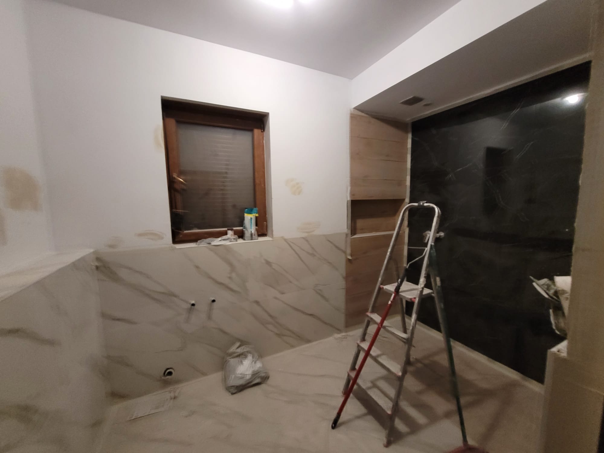 Wnętrze łazienki w trakcie prac wykończeniowych: częściowo położone płytki imitujące marmur, drabina malarska, okno z roletami i przygotowane ściany do malowania.