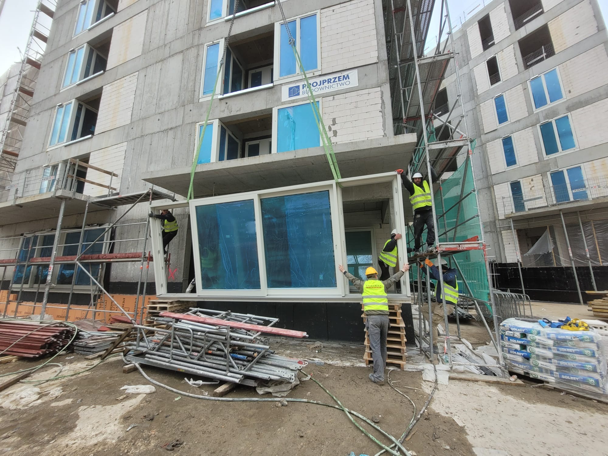 Montaż modułowej konstrukcji okiennej na placu budowy bloku mieszkalnego. Pracownicy w kamizelkach odblaskowych i kaskach. Widoczne rusztowania i materiały budowlane.