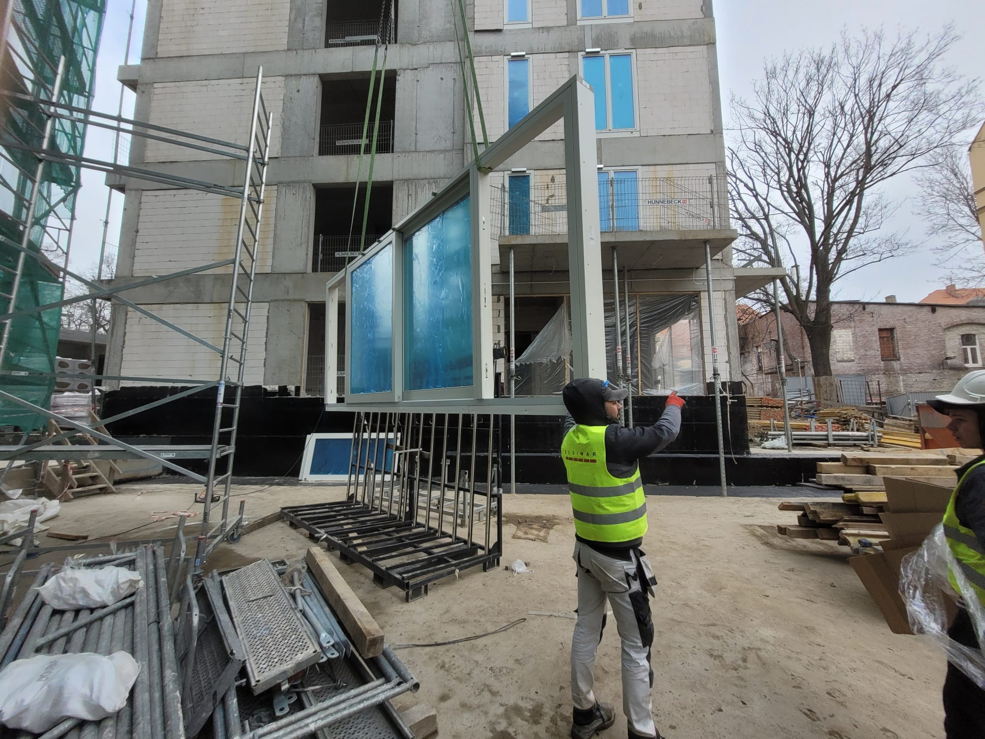Montaż okien na budowie bloku mieszkalnego. Widoczne rusztowania, pracownicy w kamizelkach odblaskowych i rama okienna podnoszona dźwigiem. Surowy stan budynku.